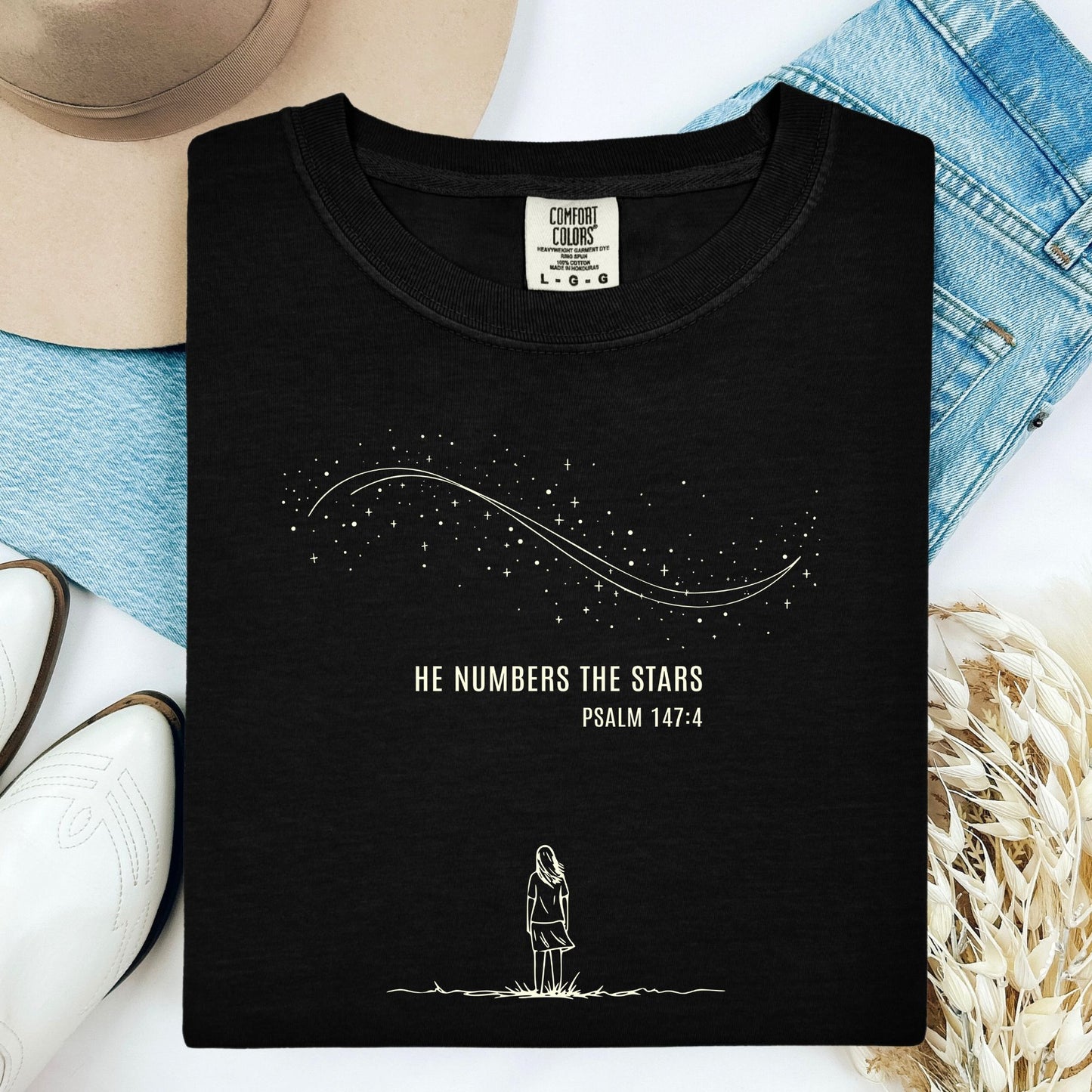 Black Comfort Colors 1717 Christian women’s T-shirt folded flat, featuring minimalist line art of a girl gazing at stars with the verse “He Numbers the Stars – Psalm 147:4.” Styled with a beige hat, denim jeans, and white boots