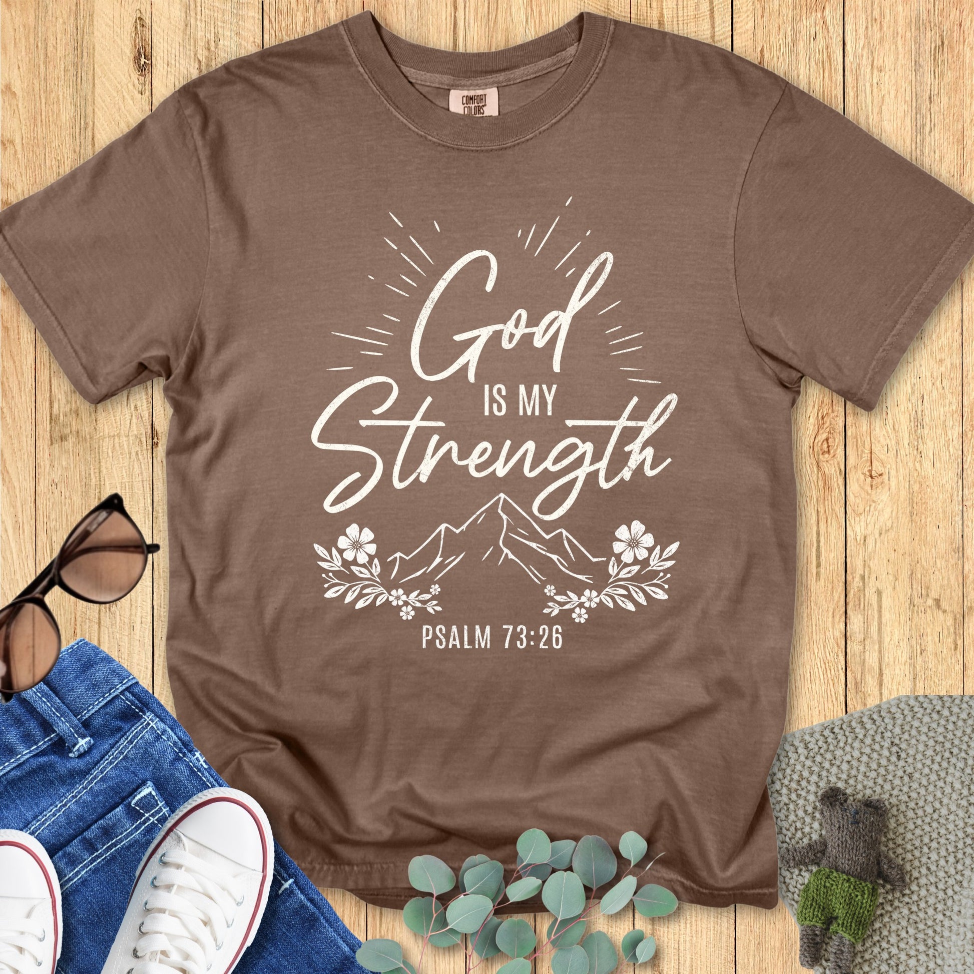 Comfort Colors 1717 espresso brown T-shirt featuring white design “God is my Strength – Psalm 73:26” with mountain, floral, and sunburst elements. Flat lay with casual lifestyle props on wood surface