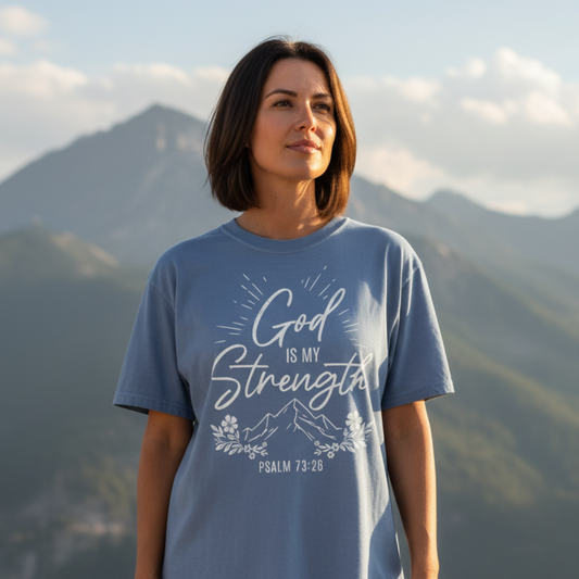 Woman standing outdoors in the mountains wearing a Comfort Colors 1717 blue jean shirt that reads โGod is my Strength โ Psalm 73:26โ with mountain, floral, and sunburst artwork