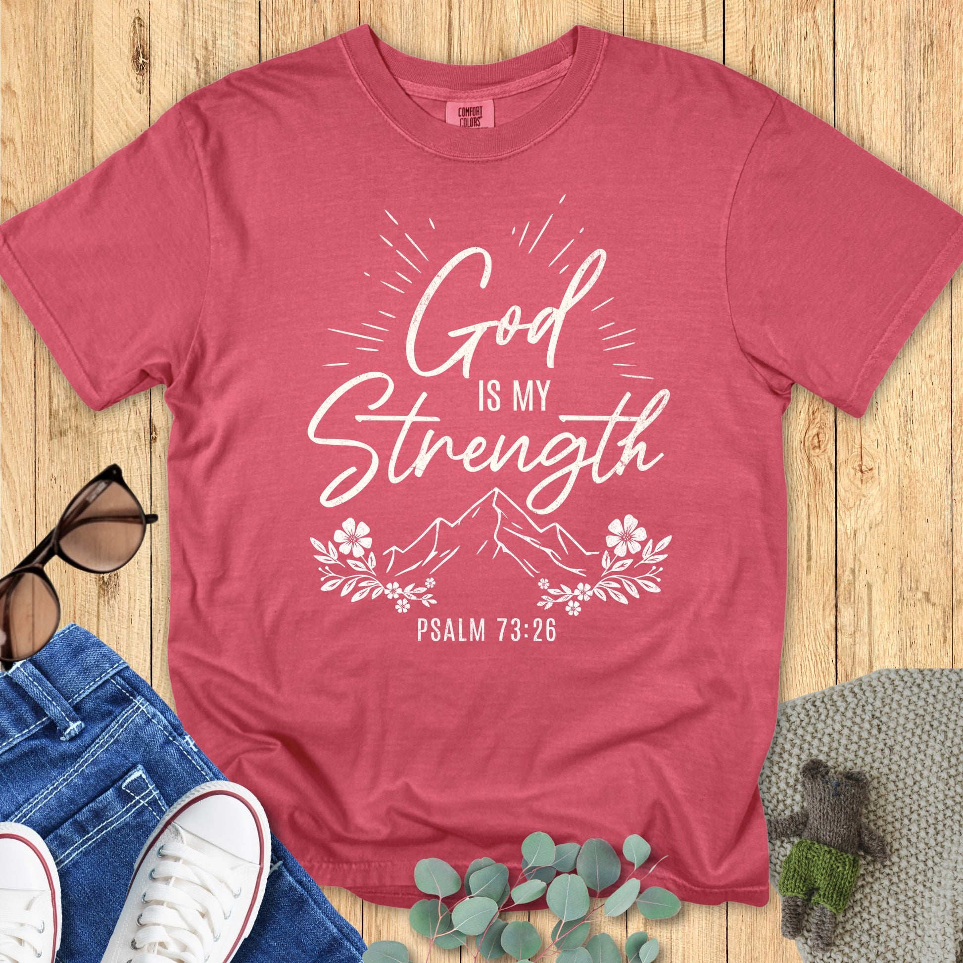 Comfort Colors 1717 crimson unisex T-shirt with white Christian design reading “God is my Strength – Psalm 73:26” in script and bold fonts, with mountain illustration, sunburst rays, and floral accents. Flat lay styled with sunglasses, denim jeans, sneakers, and greenery on a wooden background