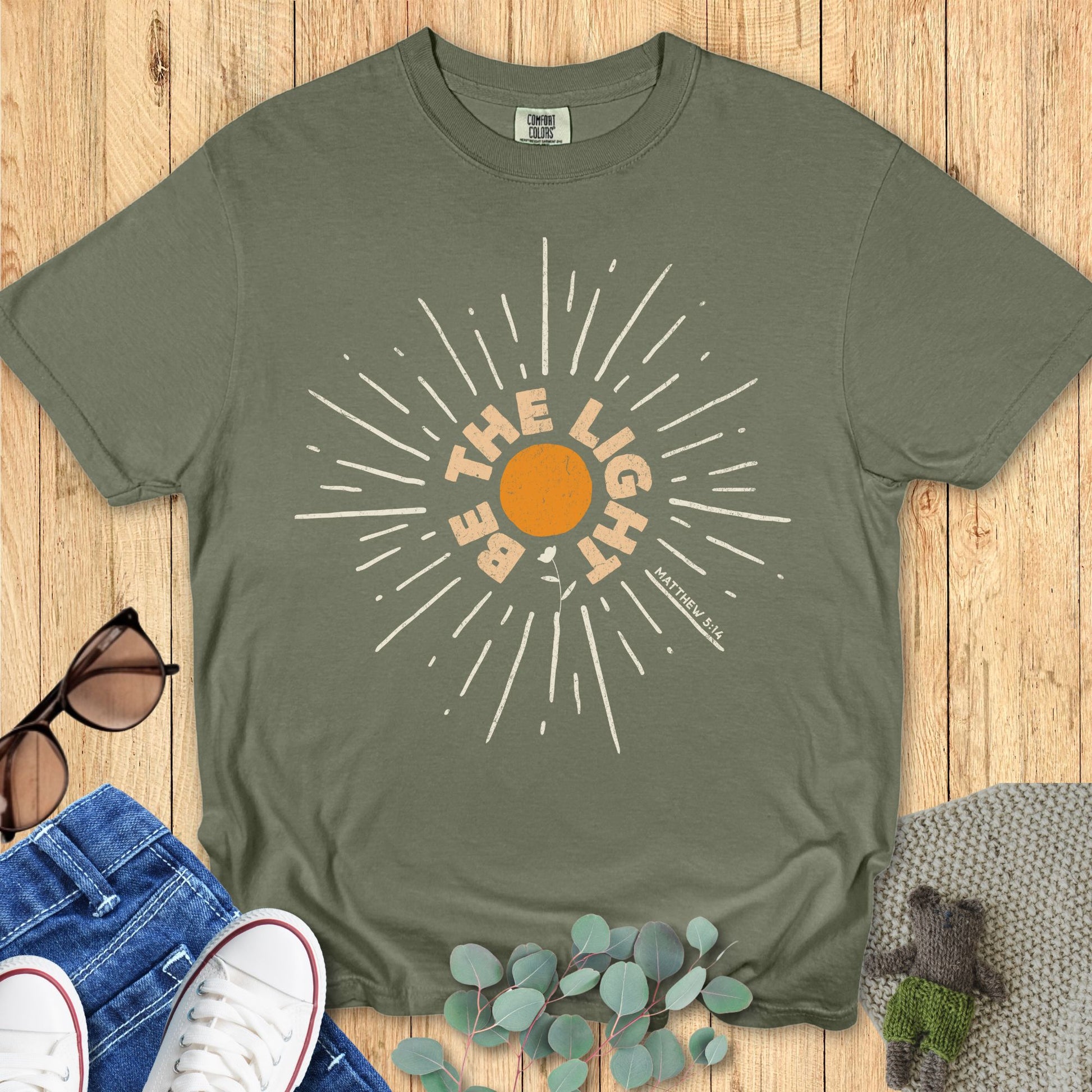 Sage green Comfort Colors 1717 Christian t-shirt laid flat on a wooden background. The print displays an orange sunburst with bold lettering: “Be The Light – Matthew 5:14,” emphasizing a message of hope and faith.