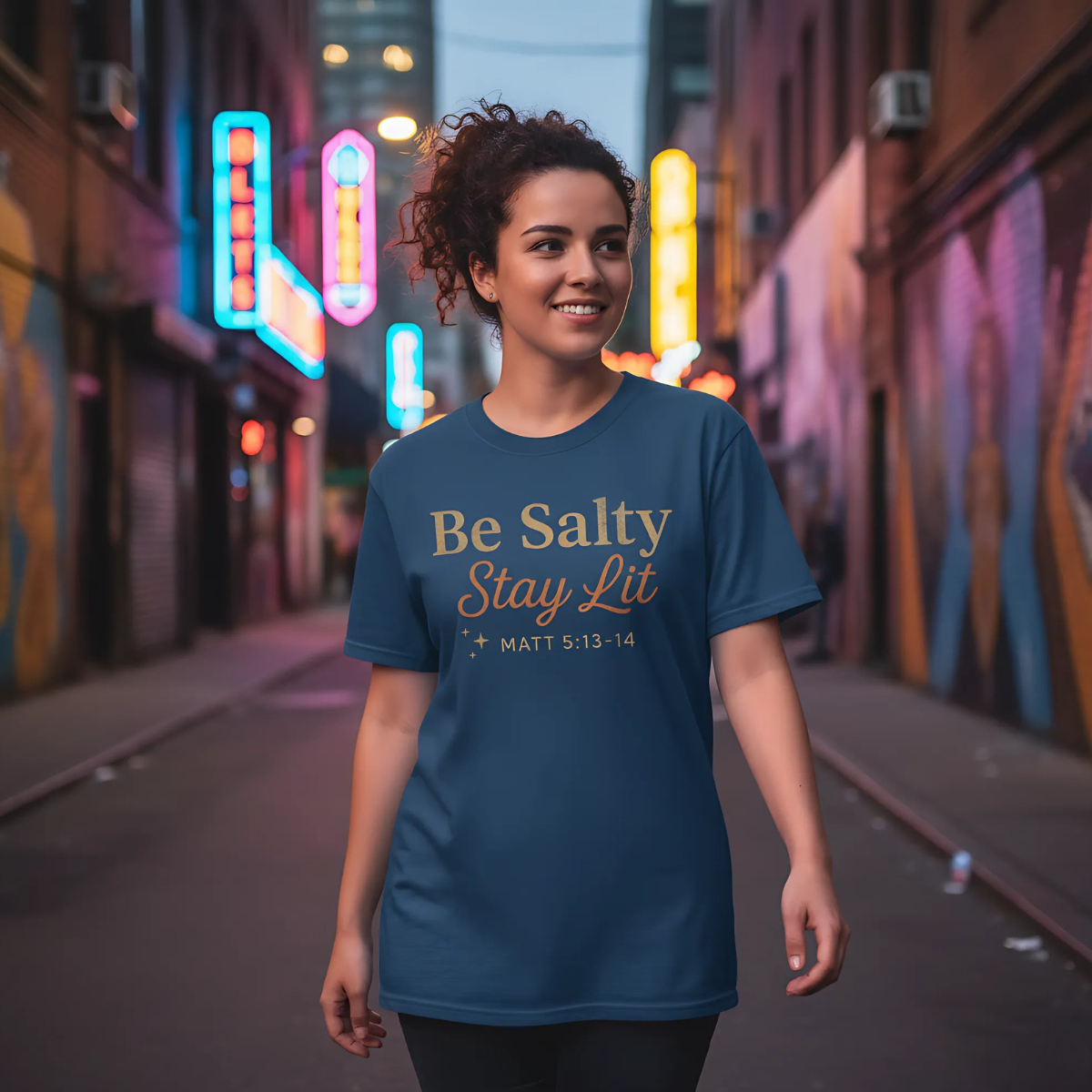 Woman walking through a city street at night wearing a blue “Be Salty Stay Lit – Matt 5:13-14” T-shirt, neon lights glowing in the background