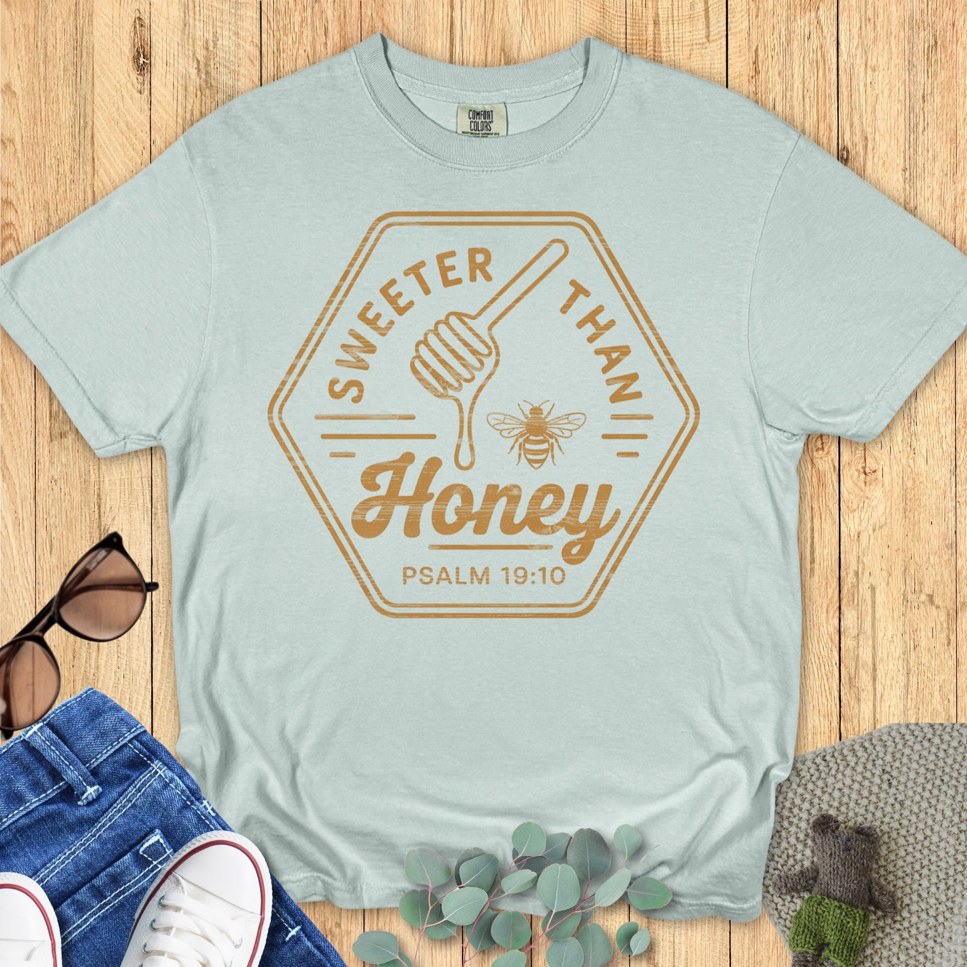 Flat lay of a mint green Comfort Colors T-shirt with “Sweeter Than Honey – Psalm 19:10” in golden brown text, honey dipper, and bee graphic. Styled with denim jeans, white sneakers, sunglasses, and eucalyptus on a wood background