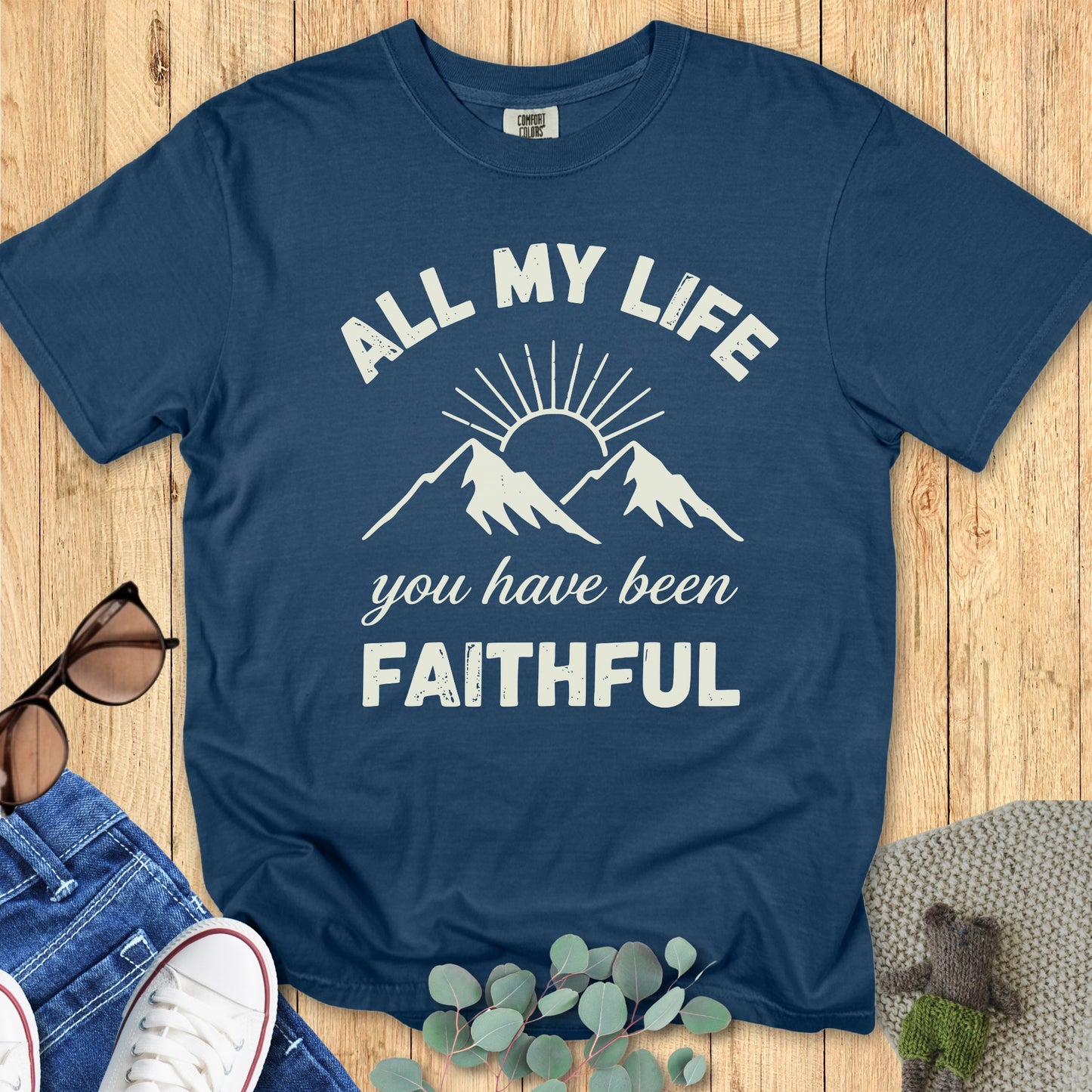 Comfort Colors 1717 midnight blue Christian T-shirt featuring mountain and sunrise graphic. Inspiring text says 'All My Life You Have Been Faithful.' Flat lay with jeans, sneakers, sunglasses, and eucalyptus leaves