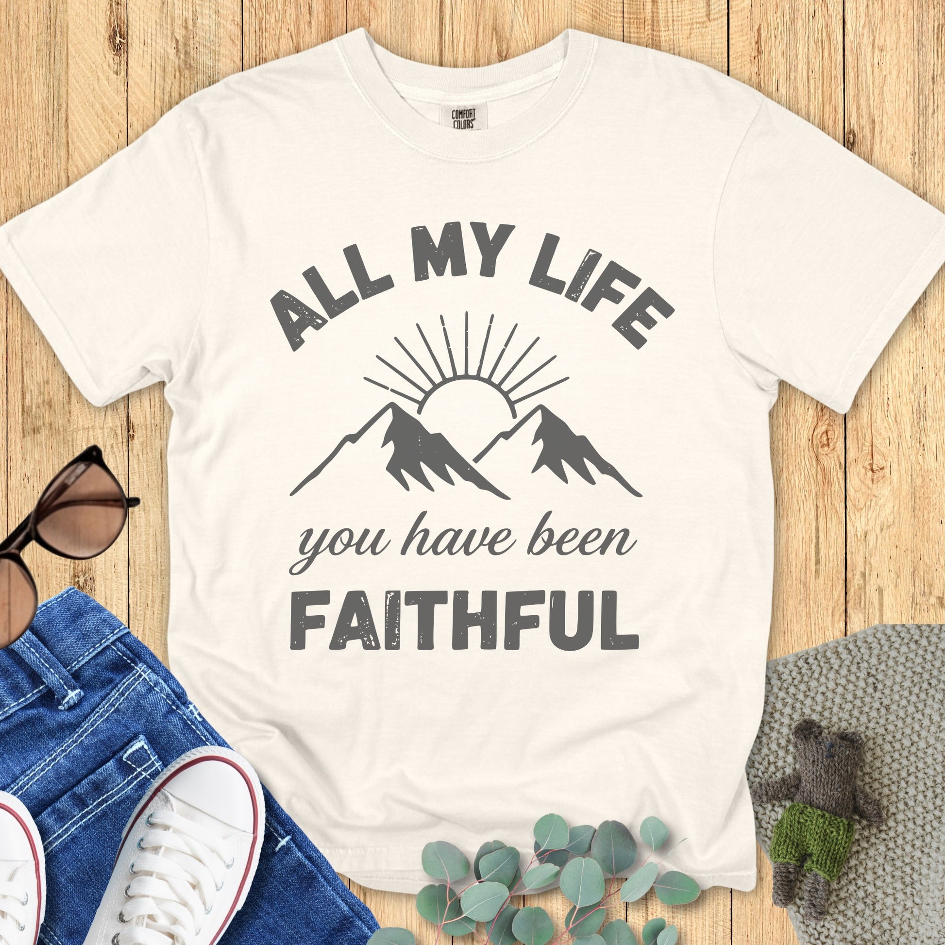 Comfort Colors 1717 ivory Christian T-shirt with minimalist mountain and sunrise graphic. Bold text reads 'All My Life You Have Been Faithful.' Flat lay styled with casual jeans, white sneakers, sunglasses, and greenery