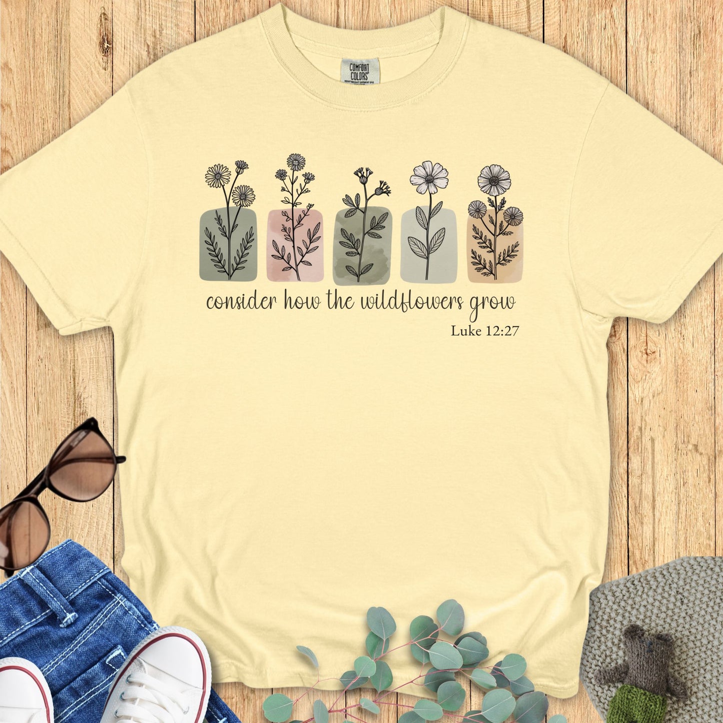 Butter Comfort Colors 1717 T-shirt mockup – Christian apparel in butter yellow, flat lay on wood background with floral graphics and Bible verse Luke 12:27, styled with sunglasses, jeans, and sneakers