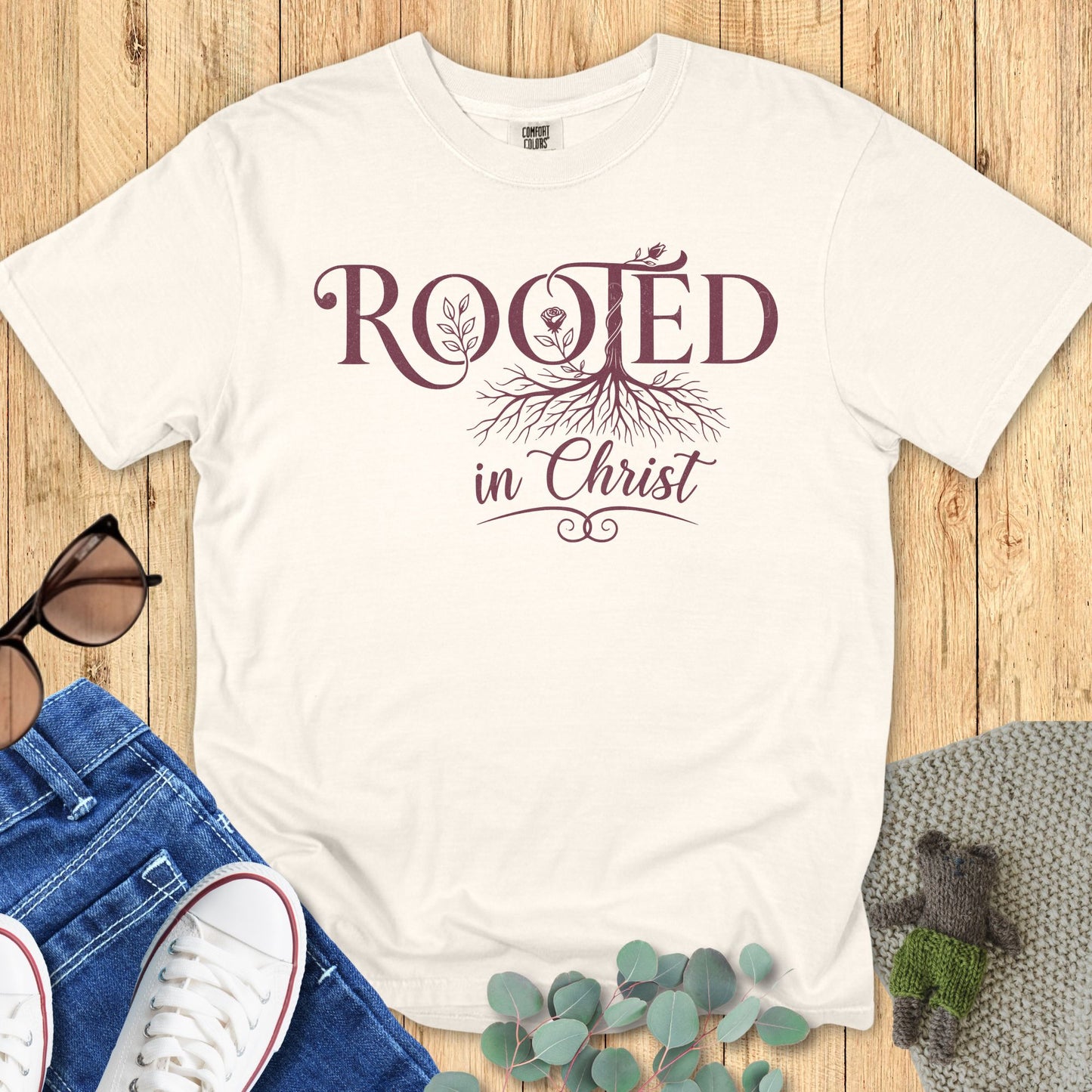 “Ivory Comfort Colors 1717 Christian T-shirt with ‘Rooted in Christ’ design in maroon lettering and tree roots graphic, styled with denim jeans and sneakers on a wooden background.”