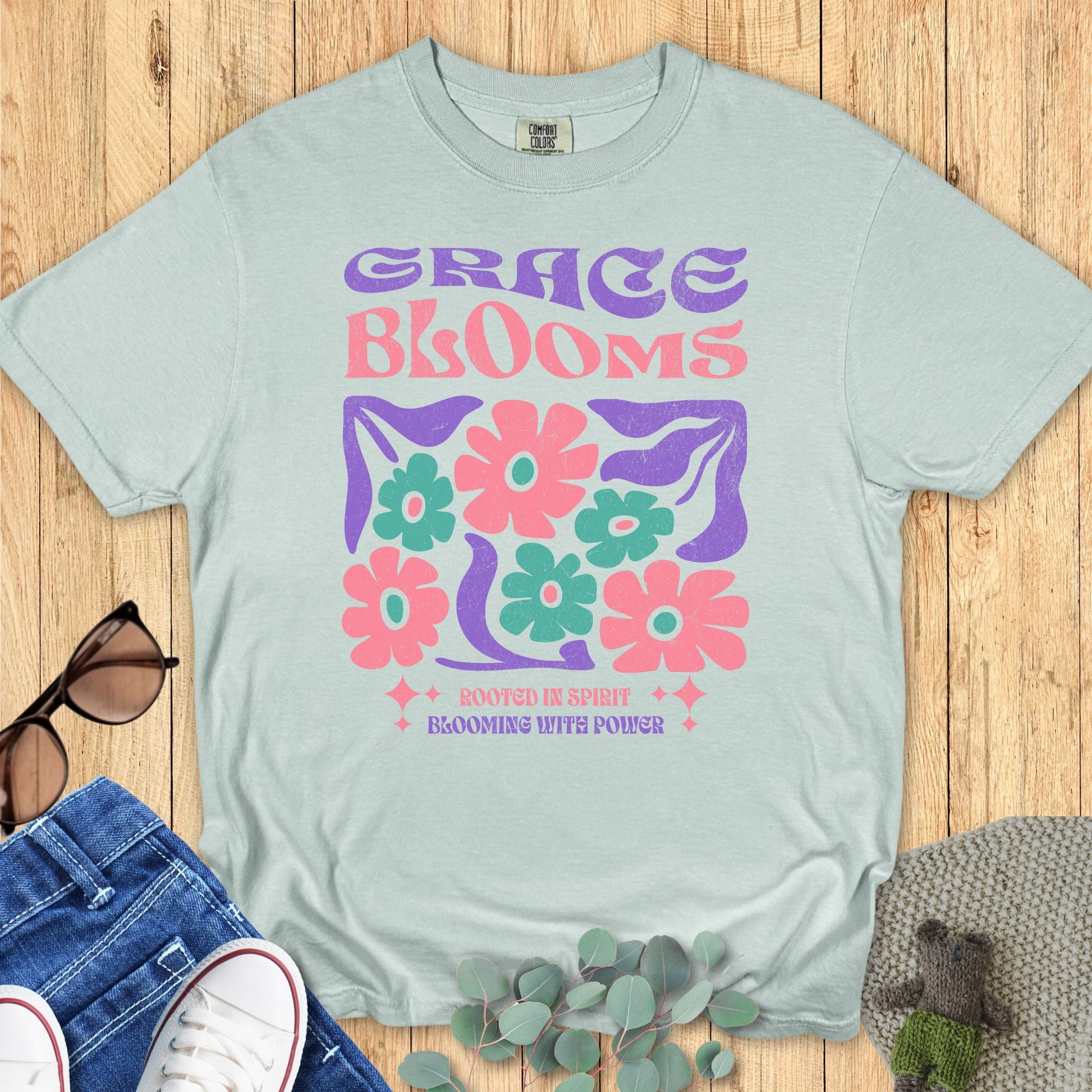 Bay green Comfort Colors 1717 Christian T-shirt with retro floral block graphic in vibrant pink, green, and purple. Reads ‘Grace Blooms’ with uplifting message ‘Rooted in Spirit, Blooming with Power.’ Flat lay Christian apparel mockup with casual styling."
