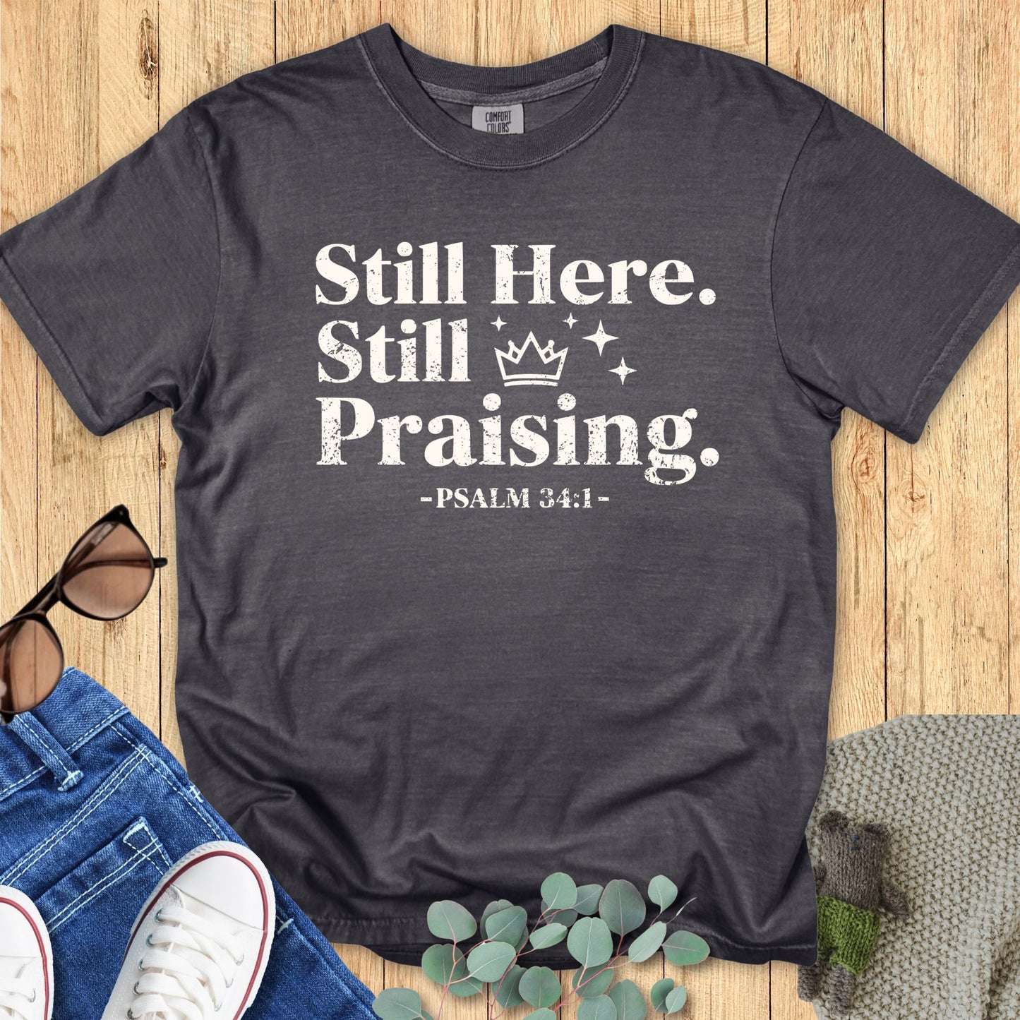 Comfort Colors 1717 Pepper gray Christian T-shirt with the phrase ‘Still Here. Still Praising. -Psalm 34:1’ in white distressed lettering, laid flat with casual accessories.