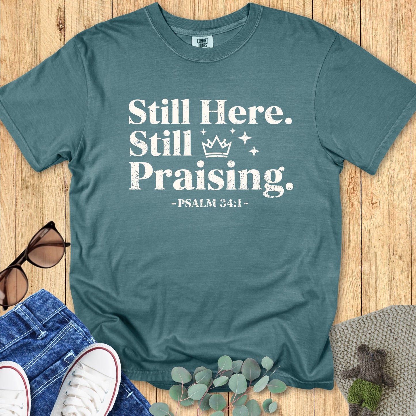 Comfort Colors 1717 Blue Spruce Christian T-shirt featuring the words ‘Still Here. Still Praising. -Psalm 34:1’ in bold white distressed font, styled with jeans, sunglasses, and eucalyptus leaves.