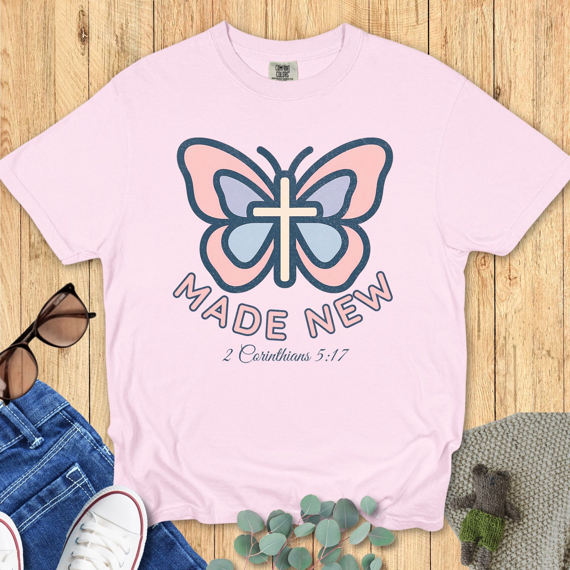 Flat lay of a Comfort Colors 1717 blossom pink Christian women’s t-shirt with butterfly and cross graphic, text “Made New – 2 Corinthians 5:17”, feminine Christian apparel style.