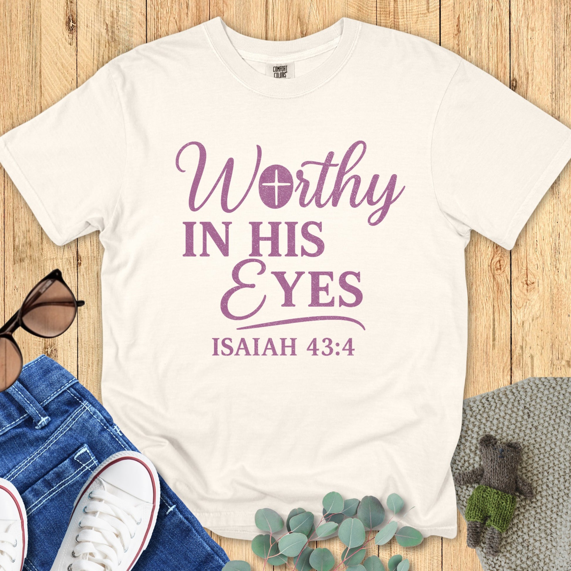 Comfort Colors 1717 ivory Christian t-shirt with the phrase 'Worthy in His Eyes – Isaiah 43:4' in purple lettering, displayed in a styled flat lay with denim jeans, sneakers, and greenery accents.