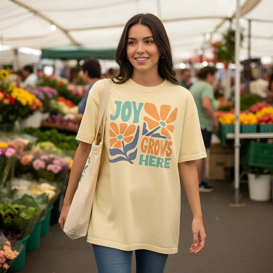 A young woman walking through a farmerโs market wearing a light yellow T-shirt with bold retro-style text that reads โJoy Grows Here,โ decorated with large orange and teal flowers