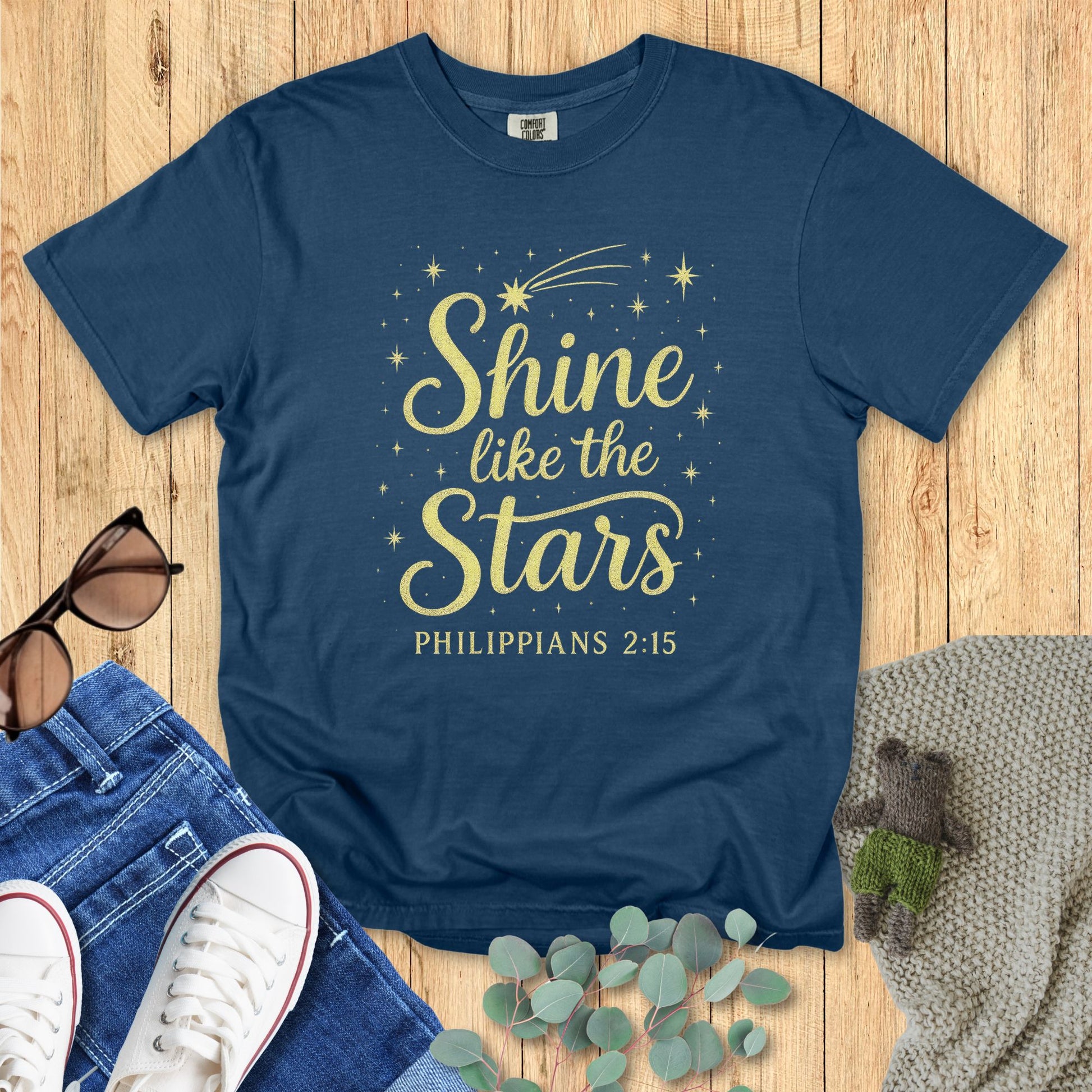 Flat lay of a midnight blue Comfort Colors 1717 T-shirt with the Christian design “Shine like the Stars – Philippians 2:15” in golden yellow, paired with jeans and casual accessories on a wooden floor.