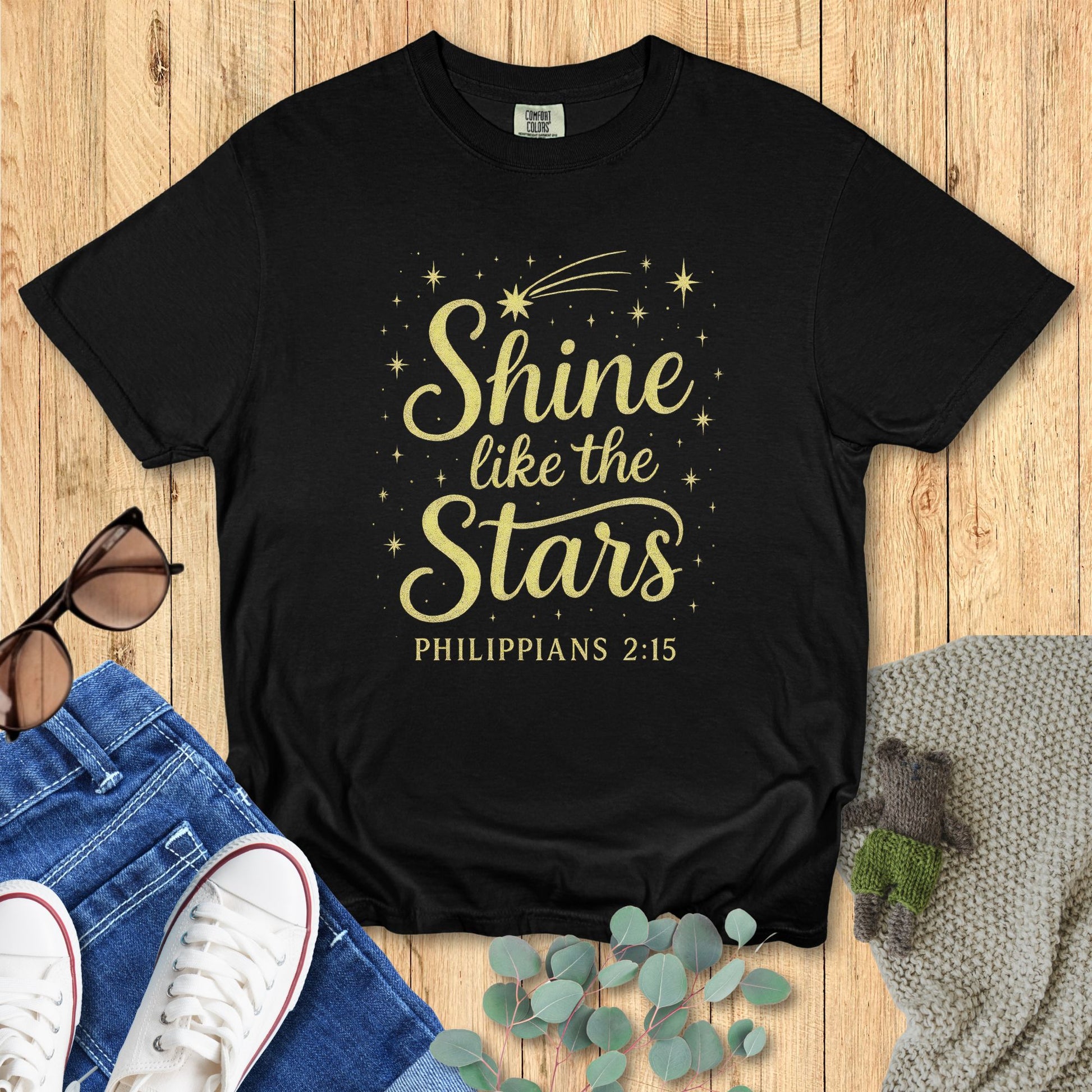 Flat lay of a black Comfort Colors 1717 T-shirt with the design “Shine like the Stars – Philippians 2:15” in gold text surrounded by stars and a comet, styled with jeans, white sneakers, and sunglasses on a wooden background.