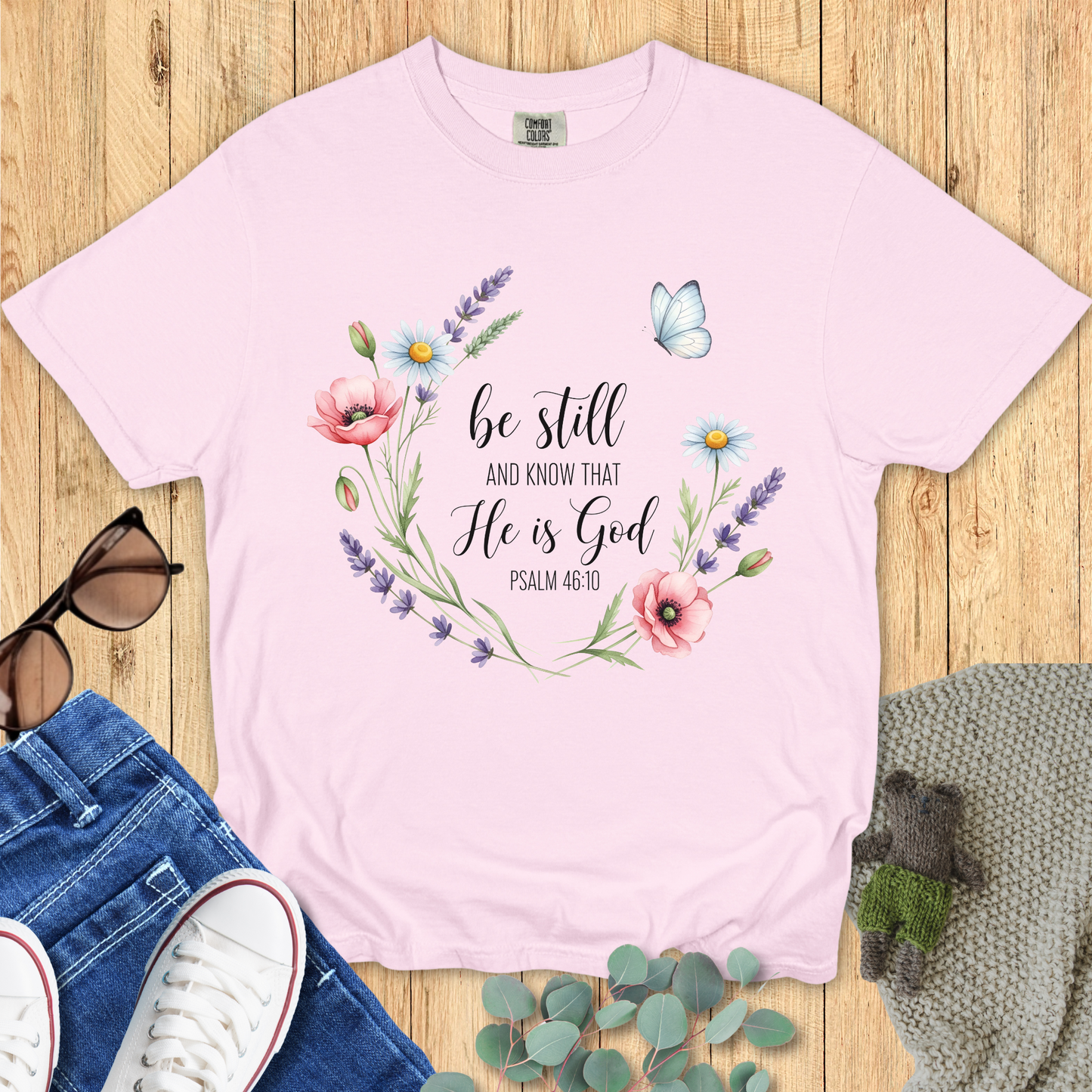 Blossom pink Comfort Colors Christian T-shirt with watercolor floral wreath and butterfly design, scripture "Be Still and Know That He Is God, Psalm 46:10." Religious inspirational tee flat lay with jeans, sneakers, and accessories.