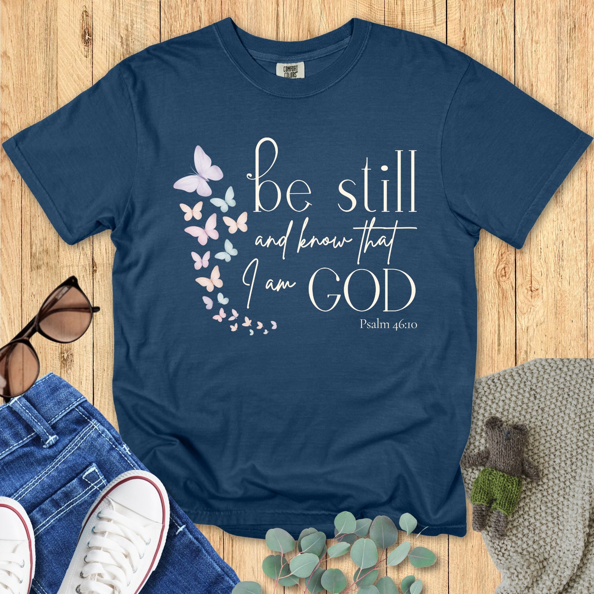 Midnight navy Comfort Colors Christian T-shirt with pastel butterfly graphic and Bible verse "Be Still and Know That I Am God, Psalm 46:10." Inspirational faith tee flat lay with jeans, sneakers, and eucalyptus leaves.