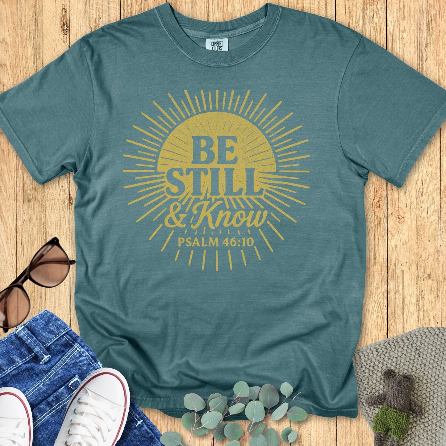 Blue Spruce Comfort Colors Christian T-shirt with golden sunburst illustration and Bible verse "Be Still & Know, Psalm 46:10." Religious inspirational tee flat lay with casual outfit accessories on wooden background.