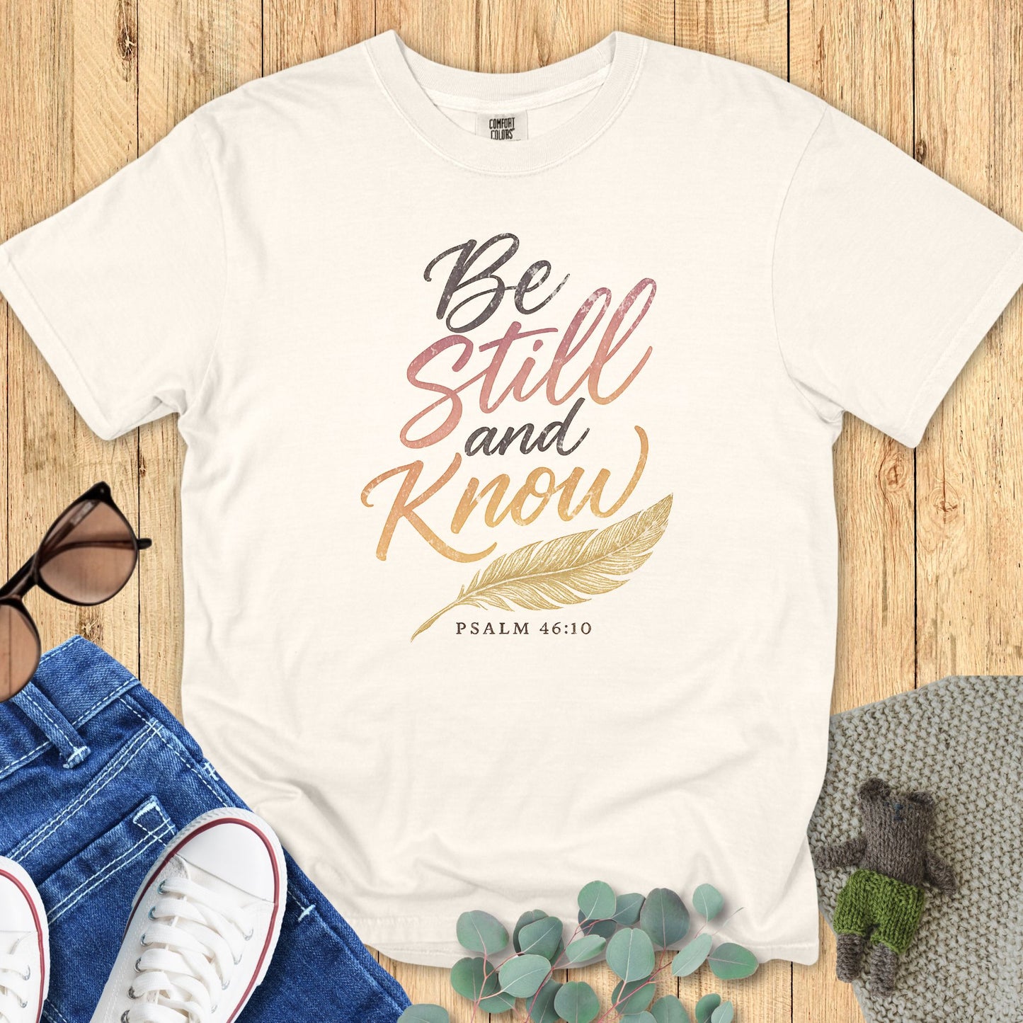 Ivory Comfort Colors Christian T-shirt with gradient script text "Be Still and Know" and gold feather graphic, Psalm 46:10. Faith-based inspirational tee flat lay with jeans, sneakers, and sunglasses on wood background.