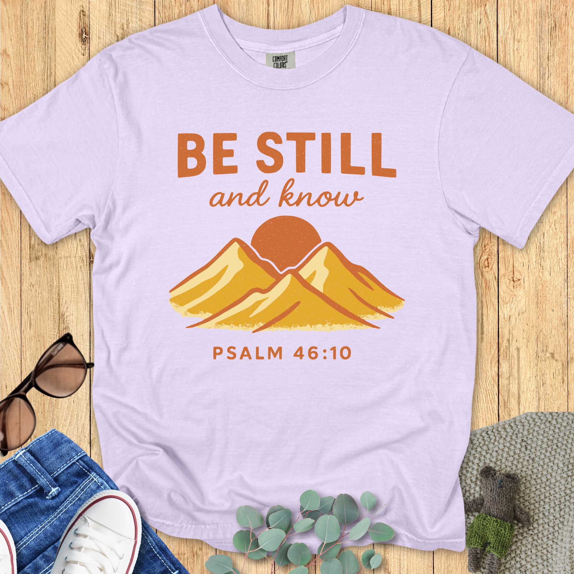 Orchid purple Comfort Colors Christian T-shirt with vintage mountain and sun design, scripture "Be Still and Know, Psalm 46:10." Faith-based graphic tee flat lay styled with denim and accessories on wood surface.