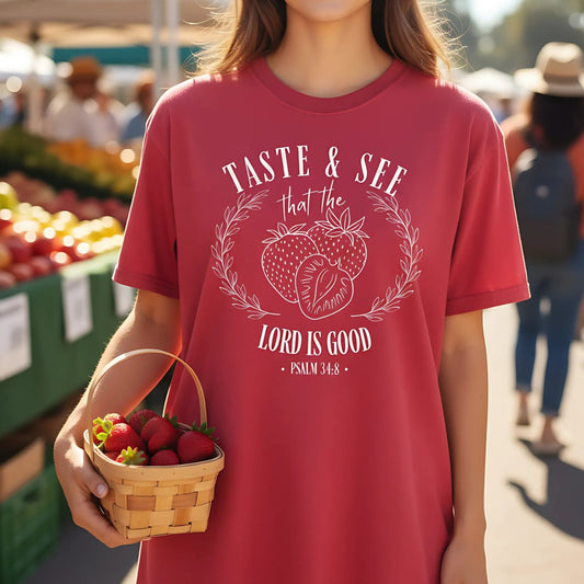 Chili Comfort Colors Christian T-shirt with strawberry wreath design and scripture "Taste & See That the Lord Is Good, Psalm 34:8." Modeled outdoors at a farmerโs market while holding a basket of fresh strawberries.