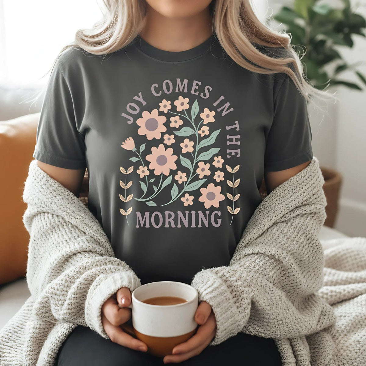 "Christian woman wearing a Comfort Colors Christian T-shirt in pepper gray with floral design and verse 'Joy Comes in the Morning.' Cozy faith-based outfit styled with cardigan and coffee cup indoors."