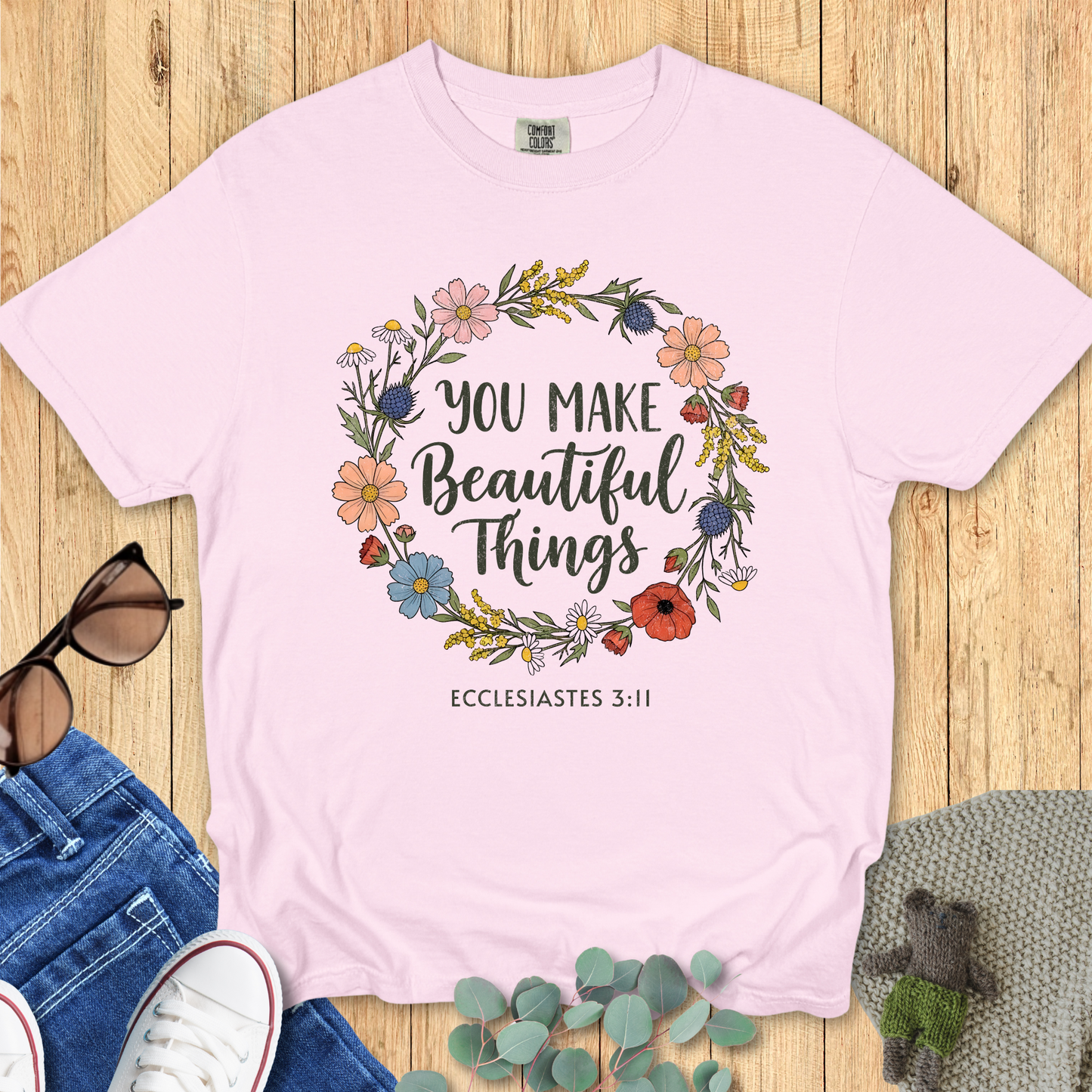 Flat lay of a blossom pink Comfort Colors Christian t-shirt featuring Ecclesiastes 3:11. The shirt design shows a hand-drawn floral wreath with the phrase “You Make Beautiful Things.” Paired with casual denim, sneakers, and eucalyptus leaves.