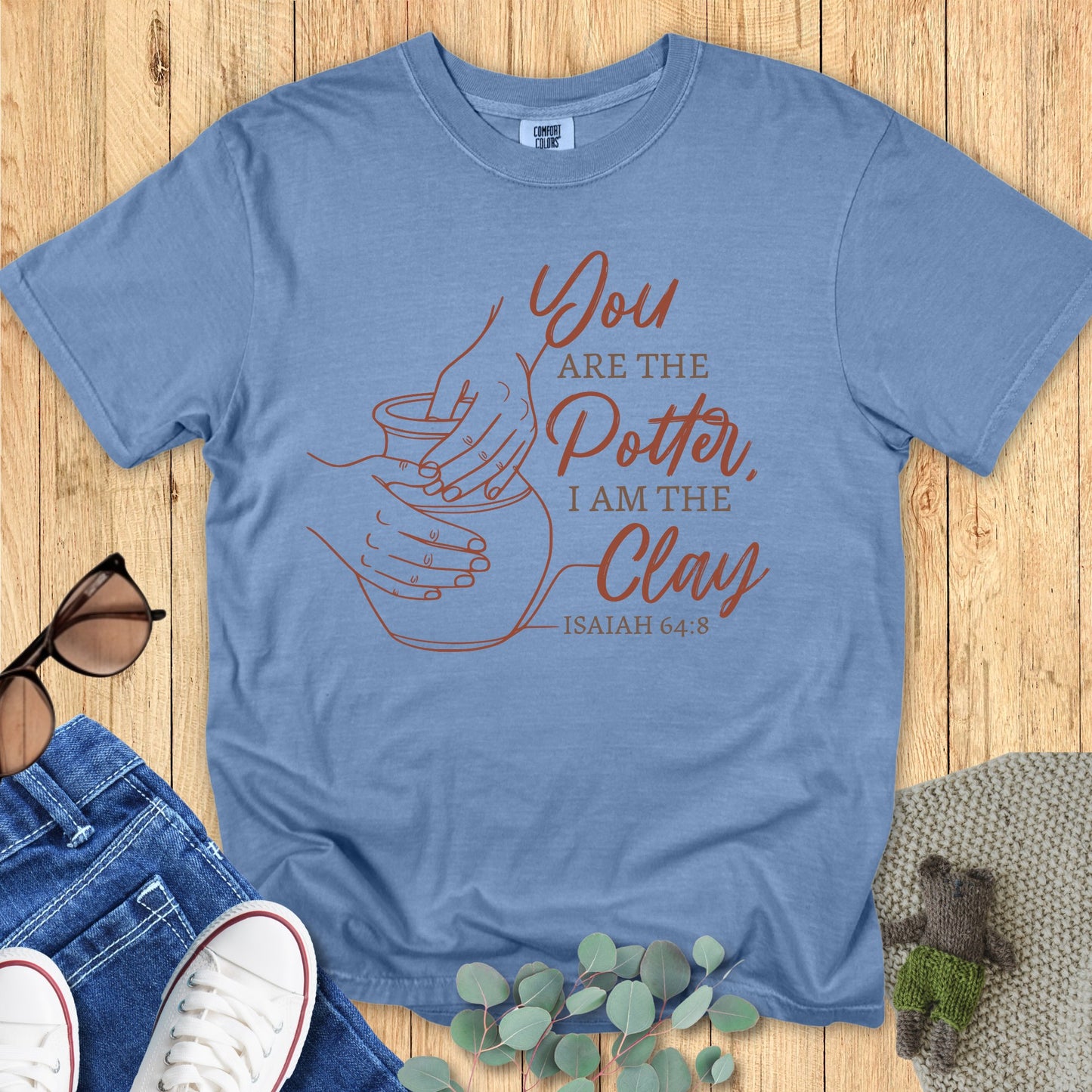 Comfort Colors Christian t-shirt in Blue Jean featuring Isaiah 64:8 verse “You are the Potter, I am the Clay” with a minimalist pottery line art design. Flat lay with jeans, sneakers, sunglasses, and eucalyptus leaves on wood background.