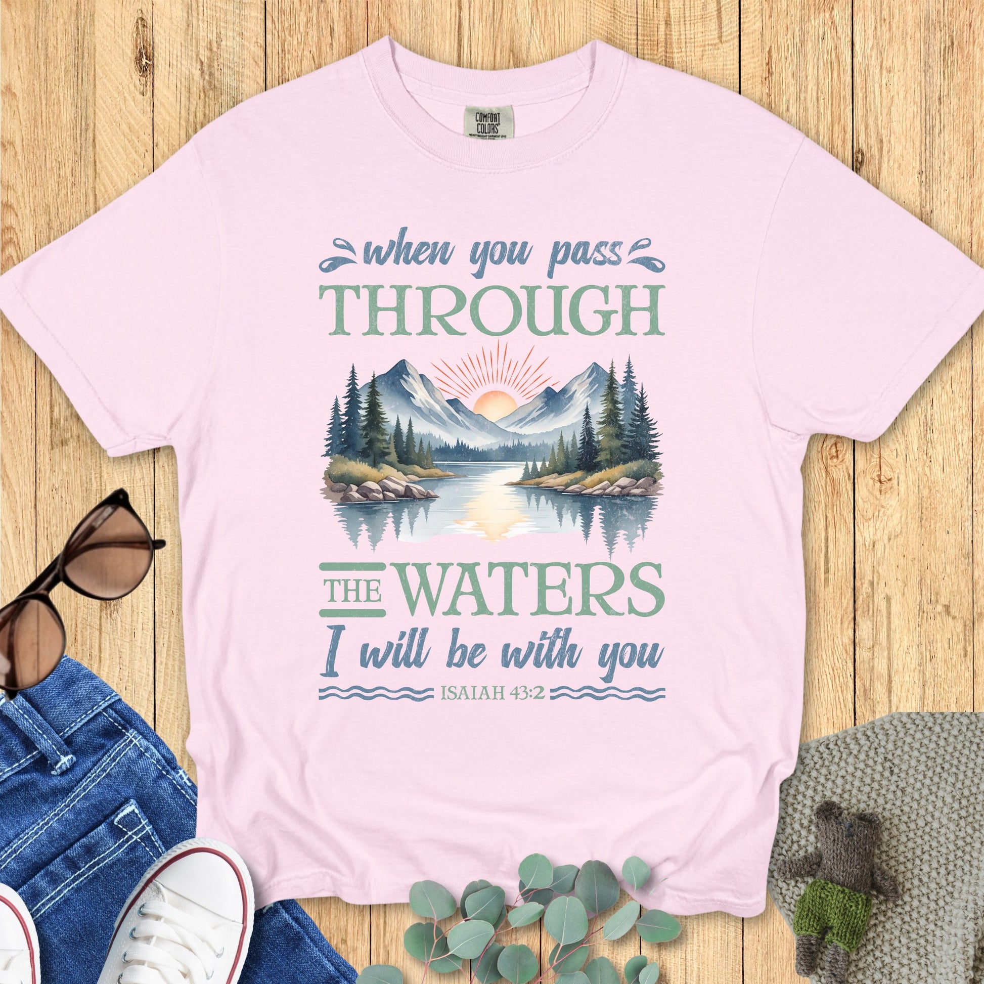 Comfort Colors Christian T-shirt in blossom pink featuring Isaiah 43:2 scripture. Illustrated graphic of mountains and water with sunrise, paired with the words “When you pass through the waters I will be with you.” Displayed flat with casual jeans, shoes, and accessories.