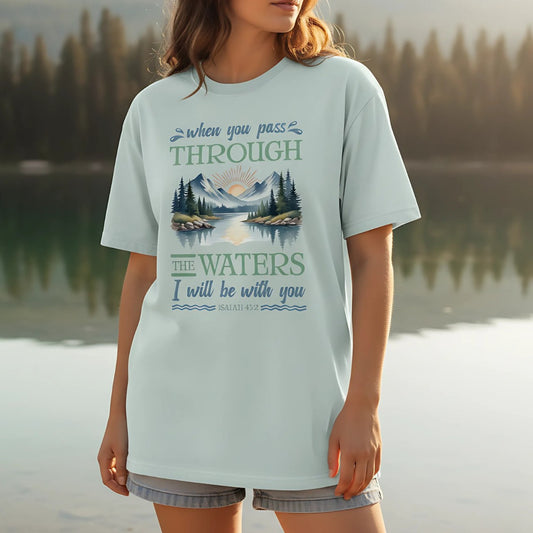 Woman wearing Comfort Colors Christian T-shirt in bay blue, standing by a peaceful lake. Shirt design includes mountain and sunrise landscape with Isaiah 43:2 verse: “When you pass through the waters I will be with you.” Styled casually with denim shorts in a natural outdoor setting.