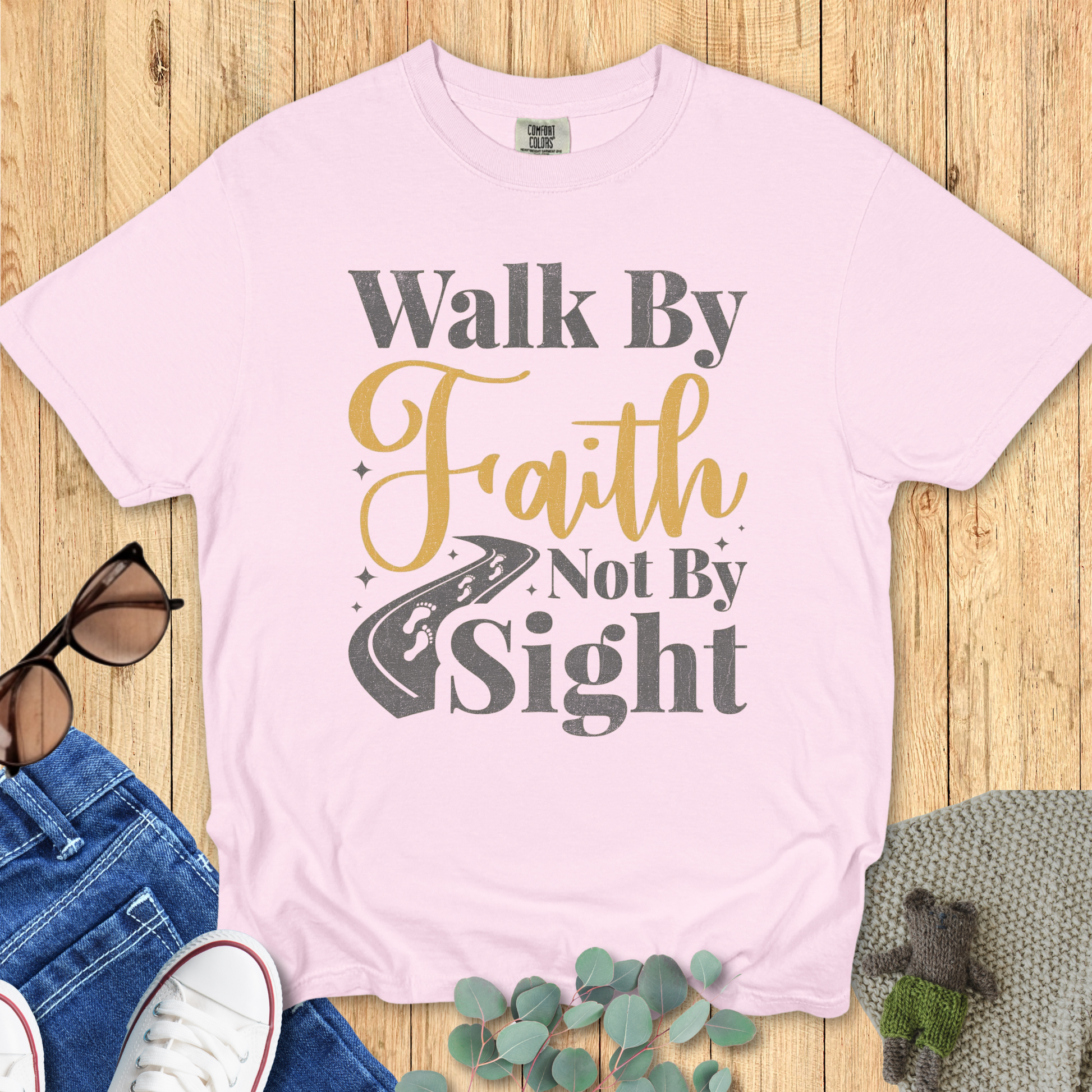 Blossom pink Comfort Colors Christian t-shirt displayed flat with the bold message 'Walk By Faith Not By Sight,' accented in gold and gray lettering with a road graphic, styled with casual flat lay accessories