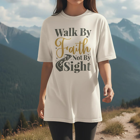 Christian woman wearing a Comfort Colors Christian t-shirt with the phrase 'Walk By Faith Not By Sight' in gold and gray lettering, styled with a road and footprints graphic, photographed outdoors on a mountain trail