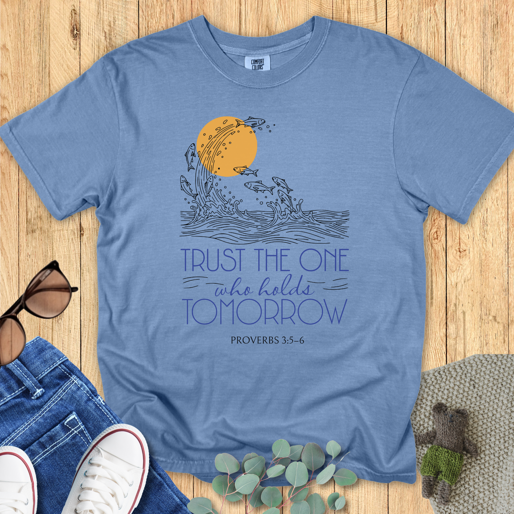 Flat lay of a blue jean Comfort Colors Christian t-shirt with Proverbs 3:5-6 scripture artwork. The print shows fish leaping from waves into the sun, symbolizing faith and trust. Shirt is styled with casual accessories on a wooden background
