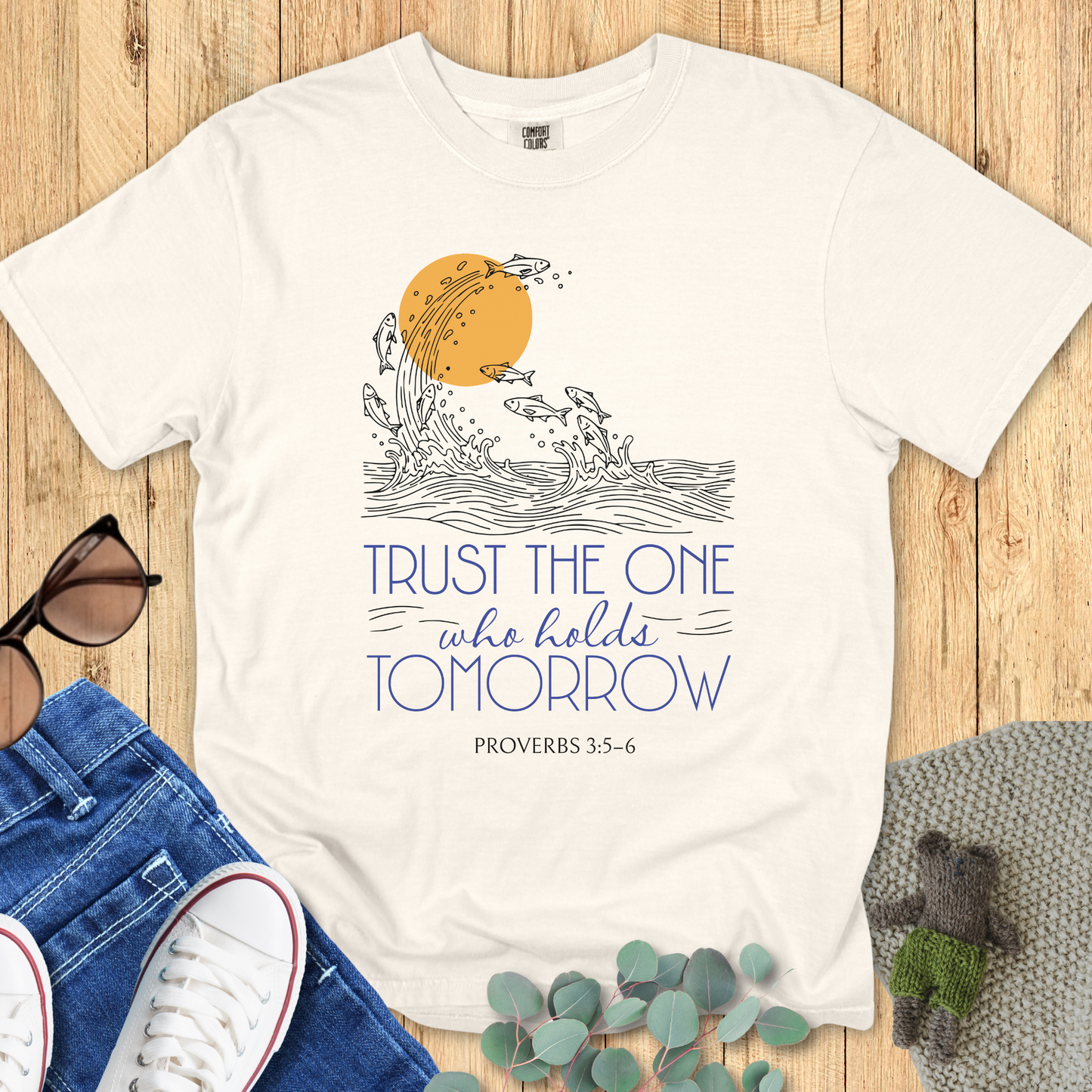 Flat lay of an ivory Comfort Colors Christian t-shirt with the Bible verse Proverbs 3:5-6 design. The shirt features a hand-drawn wave and fish graphic under a golden sun with the words “Trust the One Who Holds Tomorrow”. Styled with denim jeans, white sneakers, and sunglasses on a wooden background