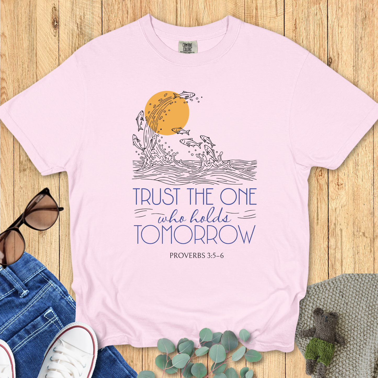 Flat lay of a blue jean Comfort Colors Christian t-shirt with Proverbs 3:5-6 scripture artwork. The print shows fish leaping from waves into the sun, symbolizing faith and trust. Shirt is styled with casual accessories on a wooden backgrounds