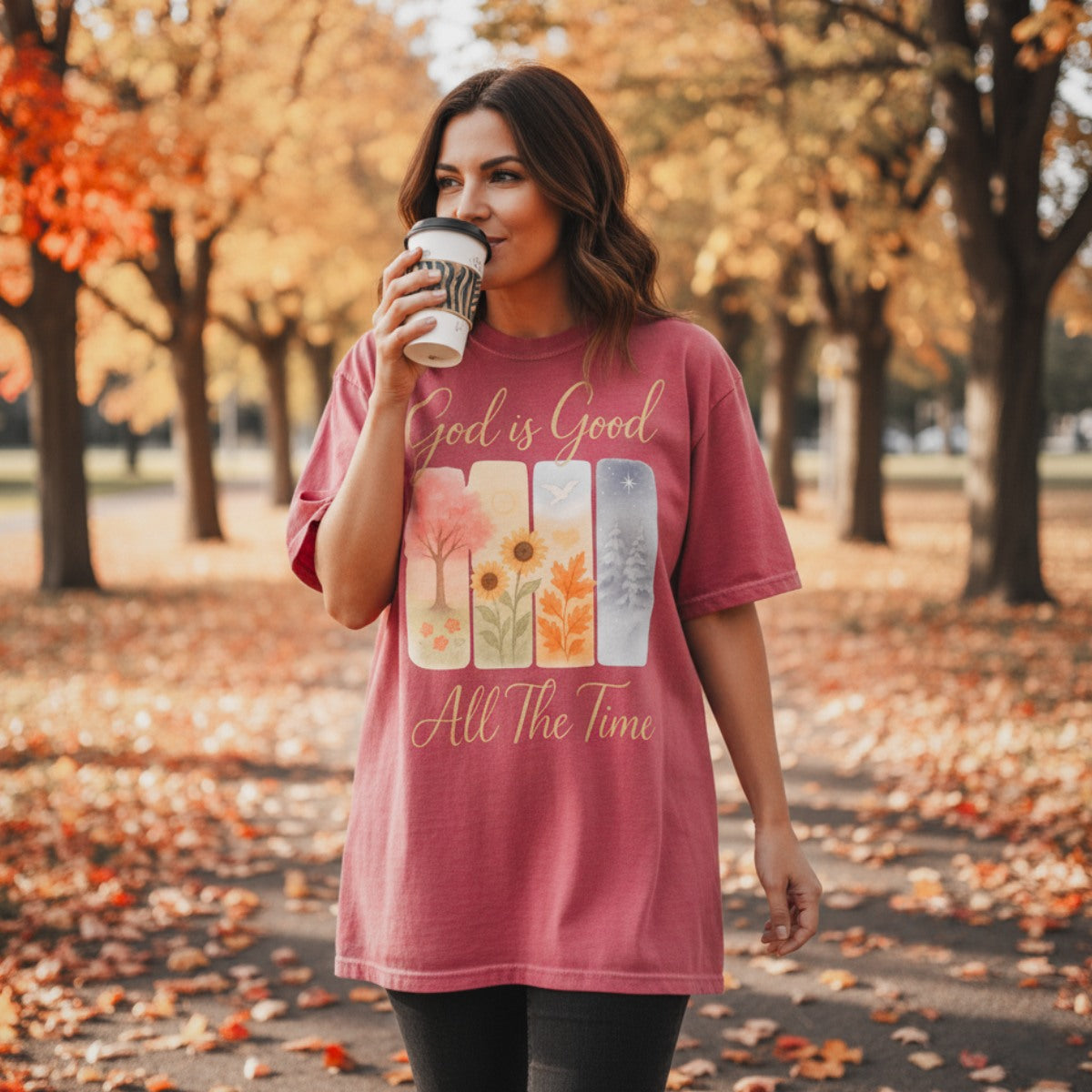 Christian women wearing a Comfort Colors 1717 crimson t-shirt with the inspirational design "God is Good All The Time." The seasonal artwork features spring, summer, fall, and winter scenes, captured in an autumn park setting with fallen leaves as she holds a coffee cup.