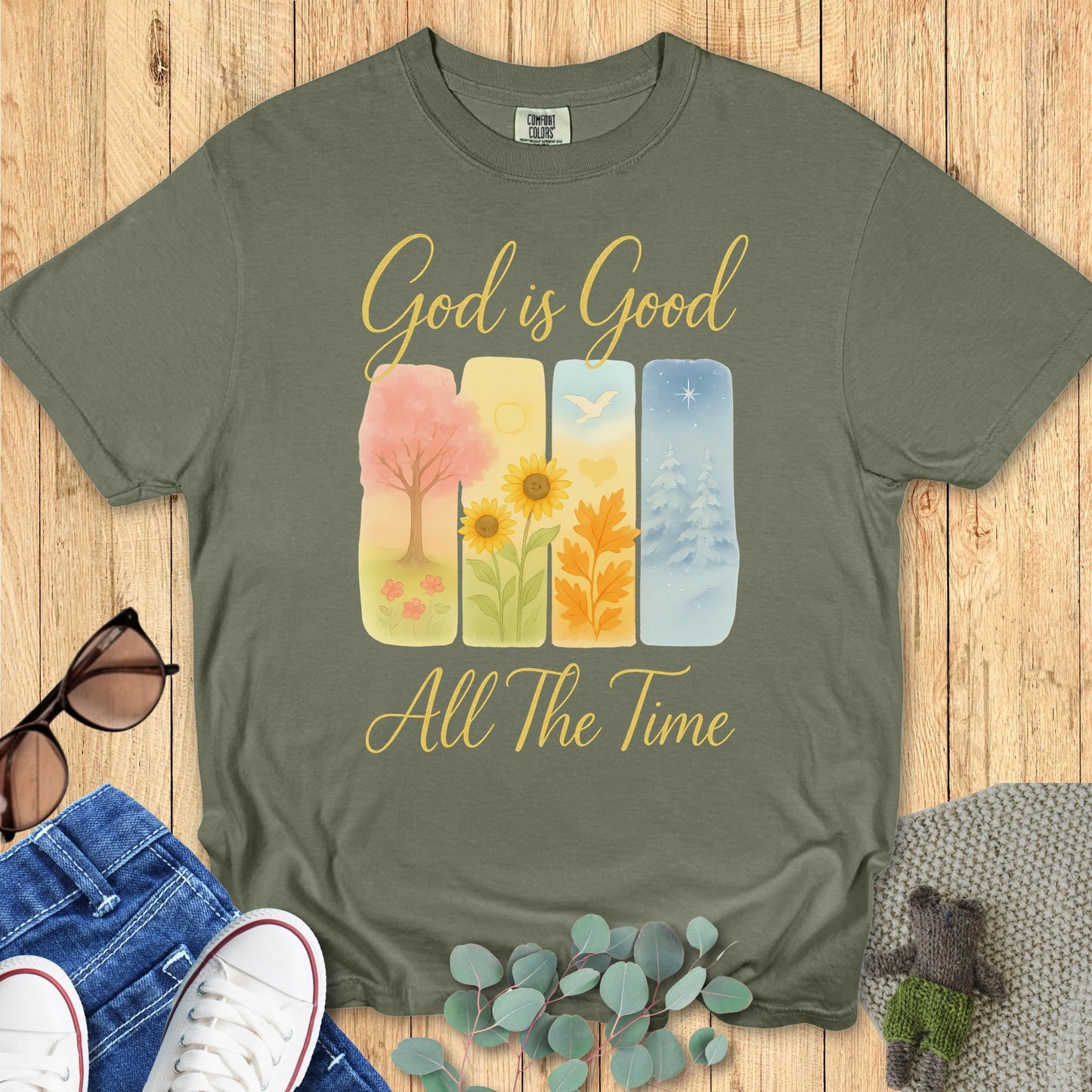 Comfort Colors 1717 Christian women t-shirt in sage green, featuring the uplifting faith design "God is Good All The Time" with four seasonal panels showing spring blossoms, summer sunflowers, autumn leaves, and winter snow. Styled in a flat lay with sunglasses, jeans, and sneakers