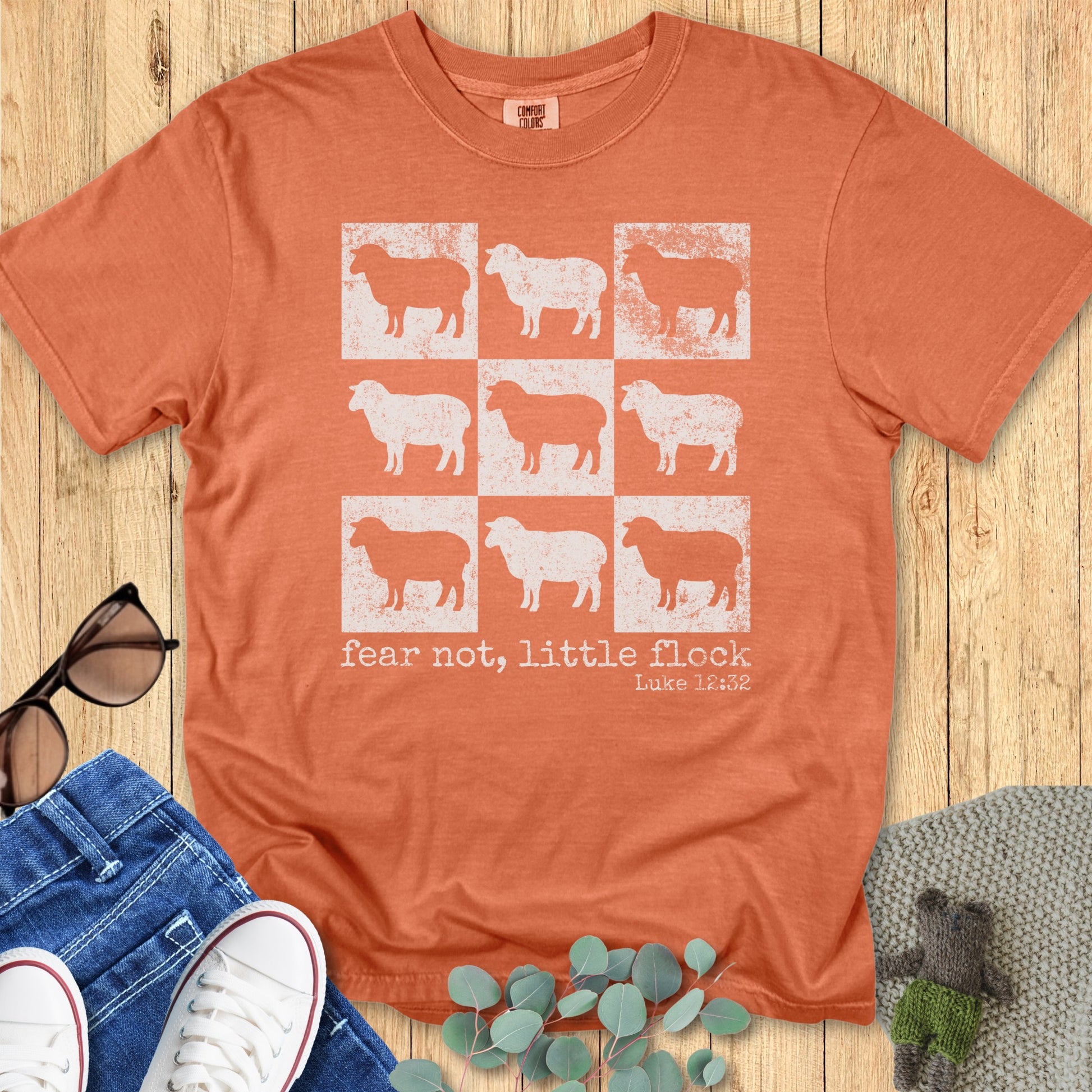 Comfort Colors 1717 Christian women t-shirt in yam orange with vintage sheep grid graphic and scripture "fear not, little flock – Luke 12:32," displayed flat lay on wooden surface with denim shorts, sunglasses, and sneakers.