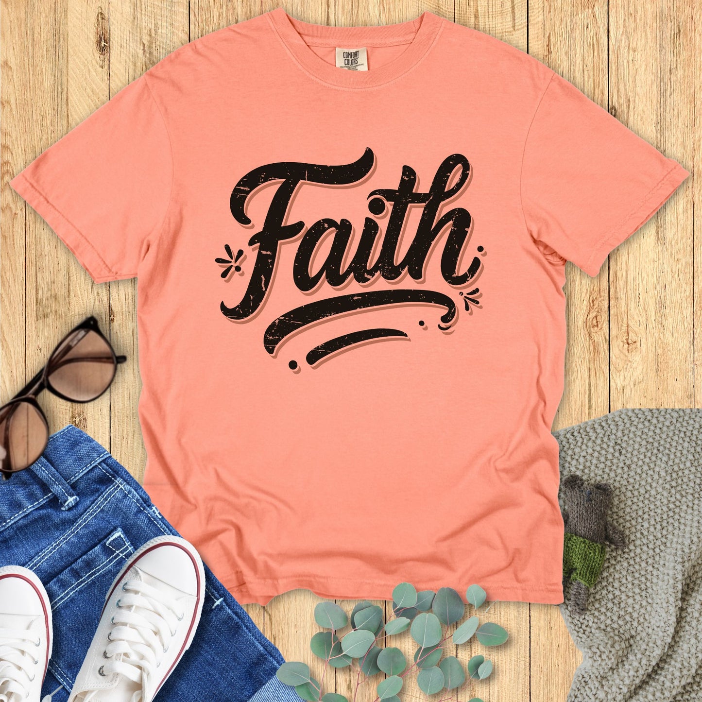 Flatlay of a Comfort Colors 1717 terracotta “Faith” t-shirt styled with jeans, white sneakers, sunglasses, and eucalyptus leaves on a wooden background. A soft vintage design from women’s scripture tees collection, perfect as a Christian mom gift idea.