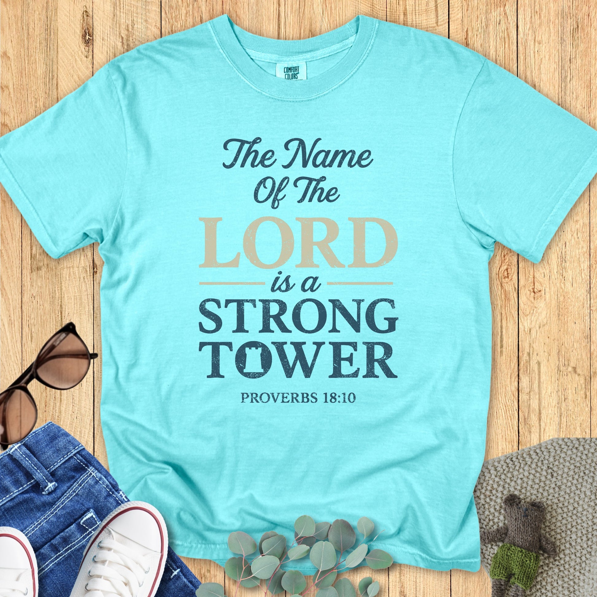 Woman standing near a peaceful lake wearing a Comfort Colors 1717 aqua “The Name of the Lord is a Strong Tower – Proverbs 18:10” t-shirt. A modern Christian shirt from faith-based apparel for women collection.