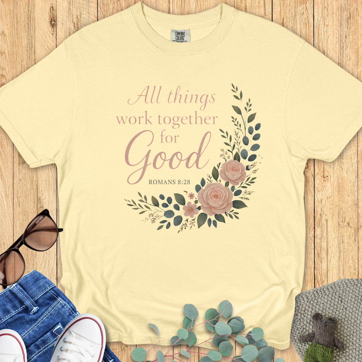 Flatlay of a Comfort Colors 1717 butter yellow “All Things Work Together for Good – Romans 8:28” t-shirt with floral artwork. A soft pastel piece from women’s scripture tees, perfect as a Christian mom gift idea.