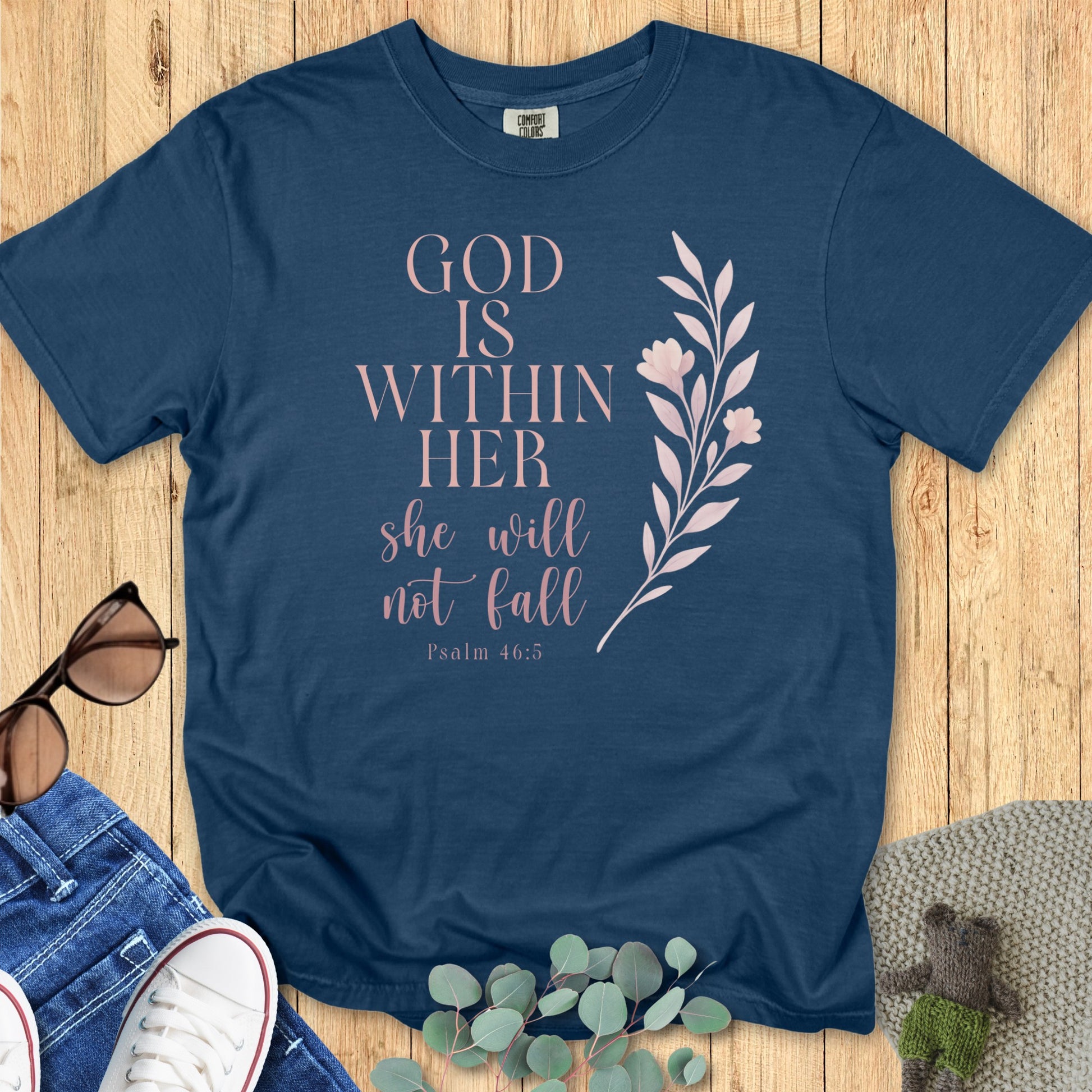 Flatlay of a Comfort Colors 1717 midnight “God Is Within Her – Psalm 46:5” t-shirt featuring elegant pink script and floral illustration. A timeless women’s scripture tee that reminds her of inner strength and faith.