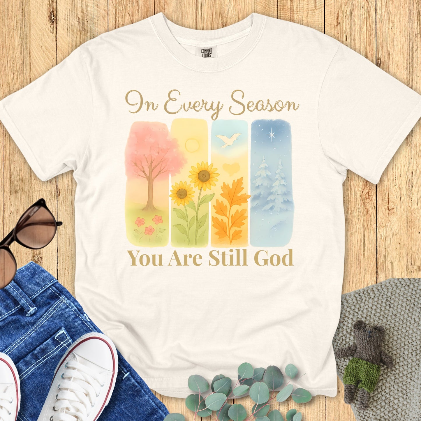 Flatlay of a Comfort Colors 1717 ivory “In Every Season You Are Still God” t-shirt with watercolor seasonal art and elegant script. A timeless women’s scripture tee and modern Christian shirt that celebrates God’s constant presence.