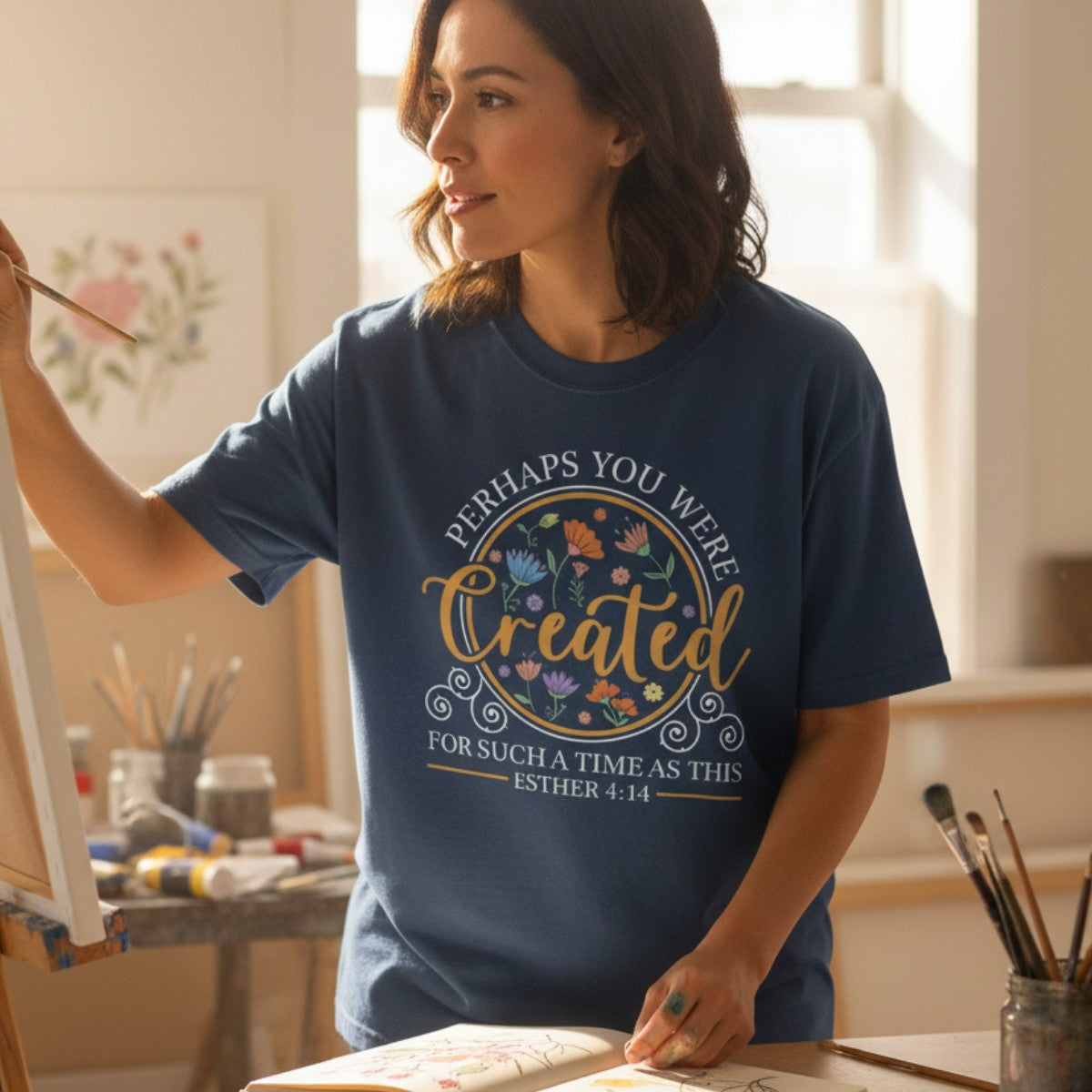 Woman painting in a sunlit studio wearing a Comfort Colors 1717 midnight “Perhaps You Were Created for Such a Time as This – Esther 4:14” t-shirt. A beautiful women’s scripture tee and piece of faith-based apparel for women that inspires creativity and confidence in God’s purpose. A thoughtful Christian mom gift idea and modern Christian shirt for everyday encouragement.