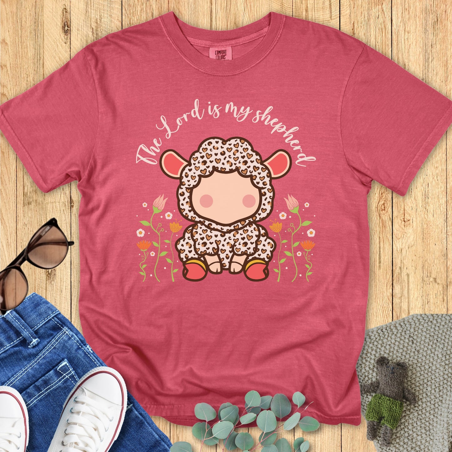 Flatlay of a Comfort Colors 1717 crimson “The Lord Is My Shepherd” t-shirt featuring an adorable cartoon lamb surrounded by flowers. A sweet women’s scripture tee and uplifting faith-based apparel for women — perfect Christian mom gift idea.