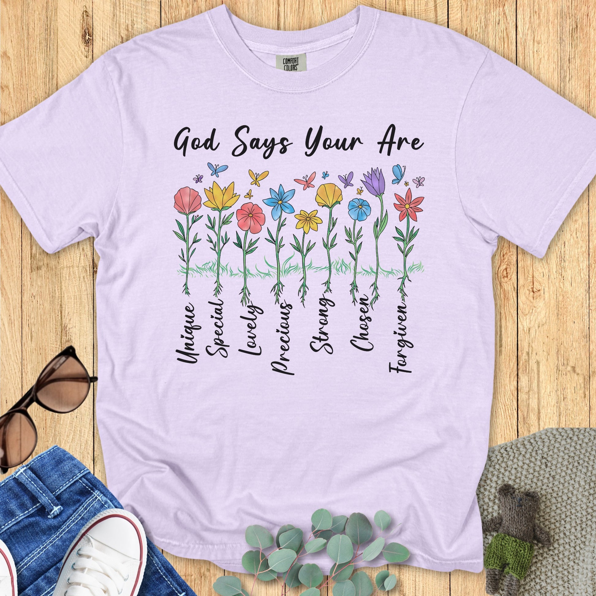 Flatlay of a Comfort Colors 1717 orchid “God Says You Are” t-shirt featuring colorful wildflowers labeled Unique, Special, Lovely, Precious, Strong, Chosen, and Forgiven. A bright and uplifting women’s scripture tee — meaningful faith-based apparel for women and a thoughtful Christian mom gift idea.
