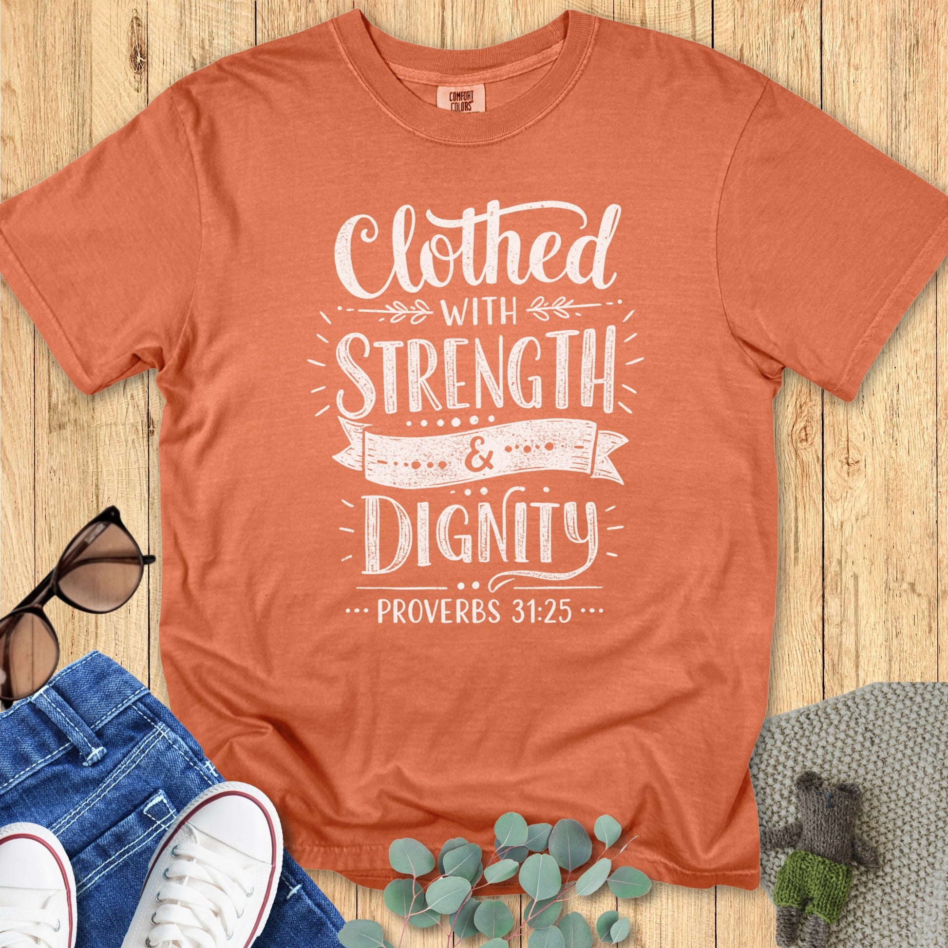 Flatlay of a Comfort Colors 1717 yam “Clothed with Strength & Dignity – Proverbs 31:25” t-shirt featuring bold white hand-lettered typography. A powerful women’s scripture tee — uplifting faith-based apparel for women and a meaningful Christian mom gift idea.