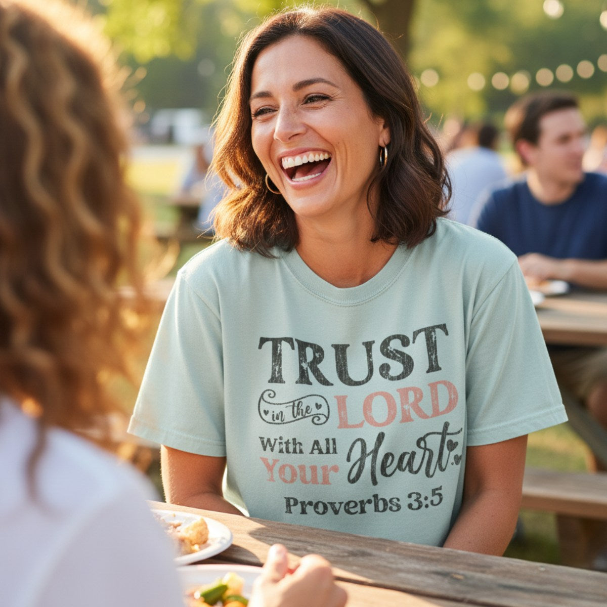 Smiling woman wearing a Comfort Colors 1717 bay “Trust in the Lord with all your heart – Proverbs 3:5” t-shirt while chatting outdoors. A joyful and uplifting women’s scripture tee — modern Christian shirt and faith-based apparel for women, perfect Christian mom gift idea.