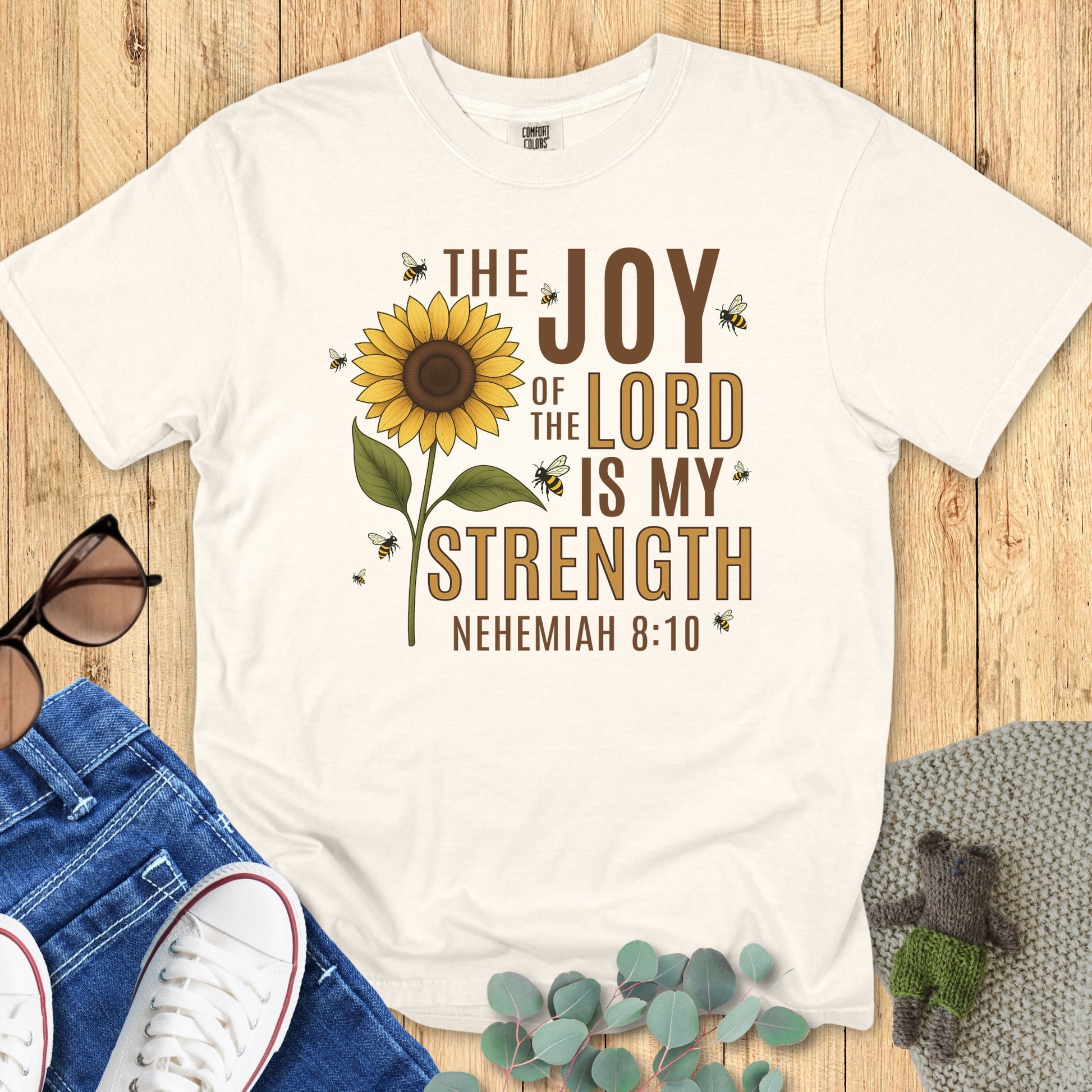 Flatlay of a Comfort Colors 1717 ivory “The Joy of the Lord Is My Strength – Nehemiah 8:10” t-shirt with sunflower and buzzing bees graphic. A bright and joyful women’s scripture tee — faith-based apparel for women and meaningful Christian mom gift idea.