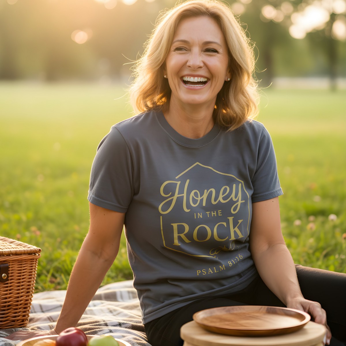 Woman laughing during a picnic wearing a Comfort Colors 1717 pepper “Honey in the Rock – Psalm 81:16” t-shirt with gold lettering. A joyful women’s scripture tee — stylish faith-based apparel for women and meaningful Christian mom gift idea inspired by Psalm 81:16.